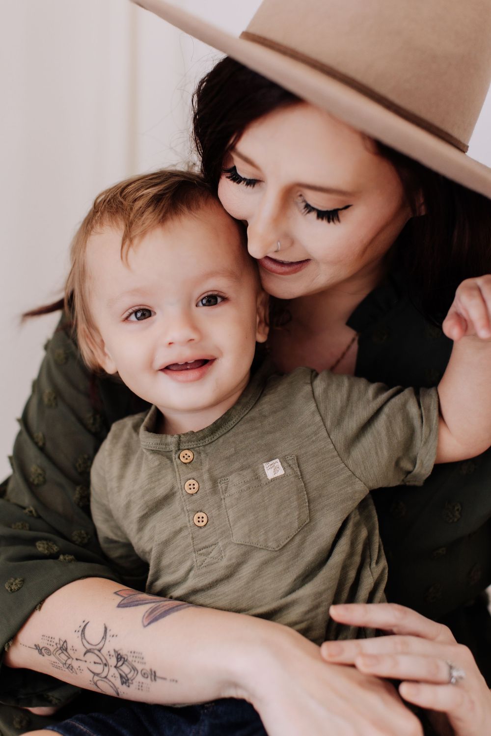 A parent wearing a beige hat shares a tender moment with their smiling baby dressed in olive green.