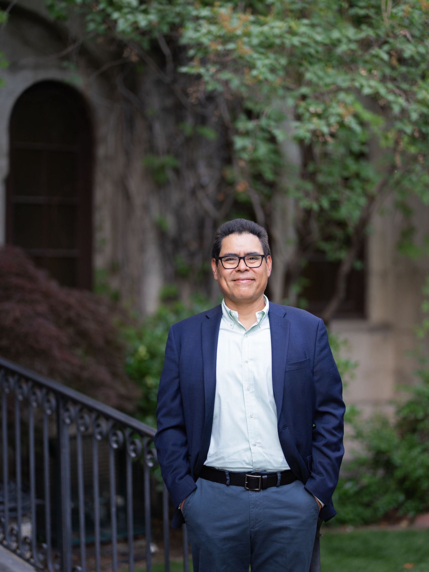 Professional wearing a navy blazer and white dress shirt poses confidently against a wall with green ivy foliage.