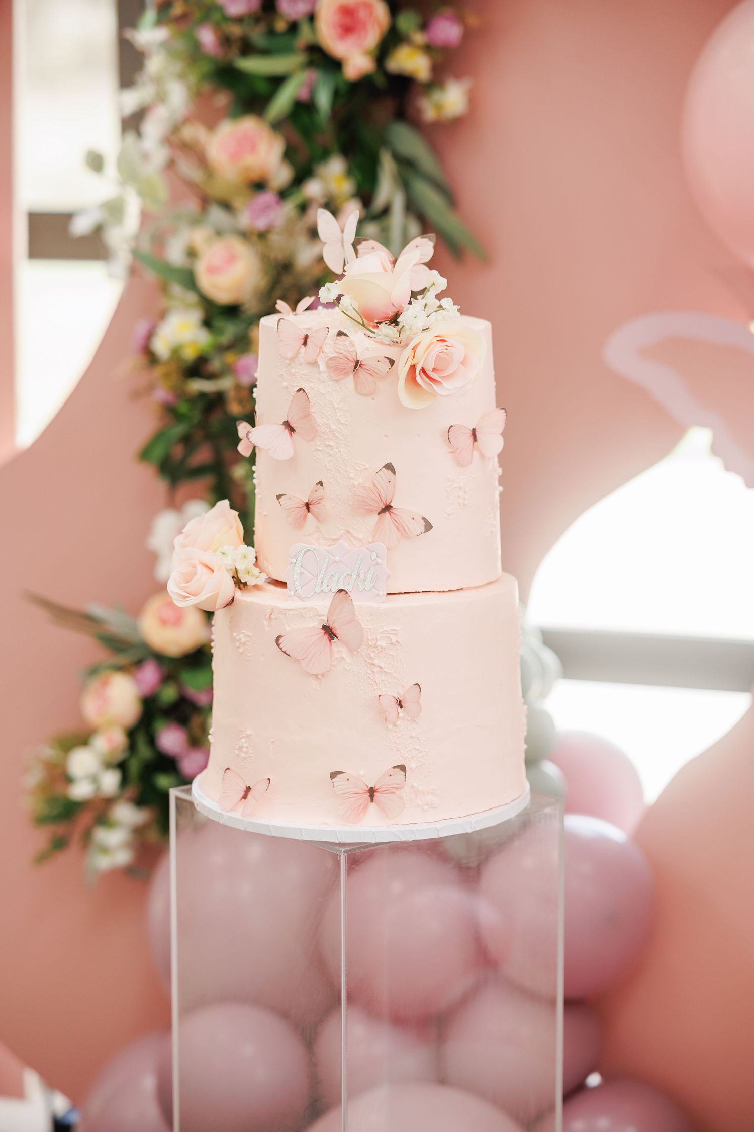 Elegant two-tier pink birthday cake decorated with delicate sugar flowers and butterflies on clear stand with pink balloon backdrop.