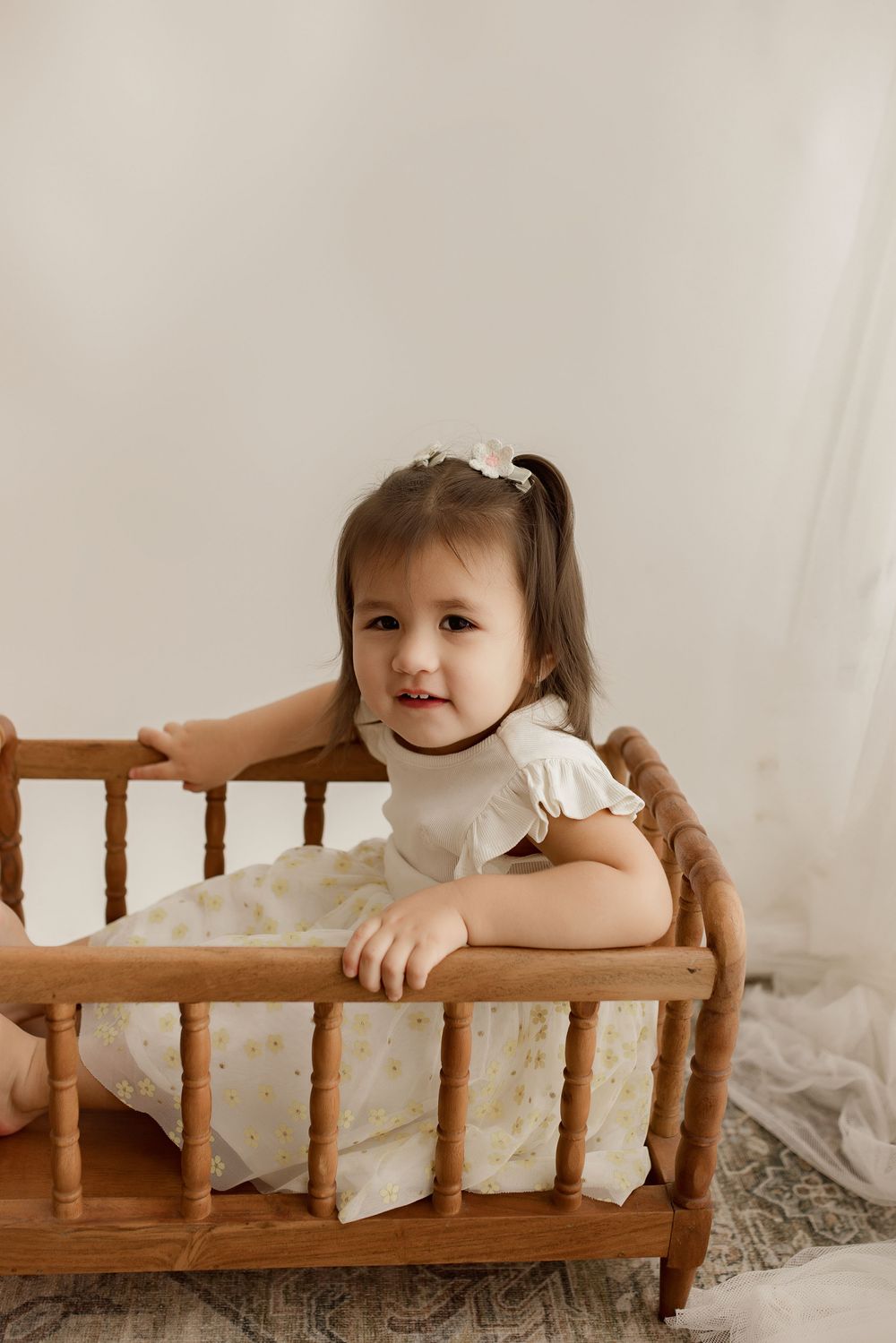 A toddler in a white dress leans against a vintage wooden doll crib with dreamy neutral-toned backdrop.