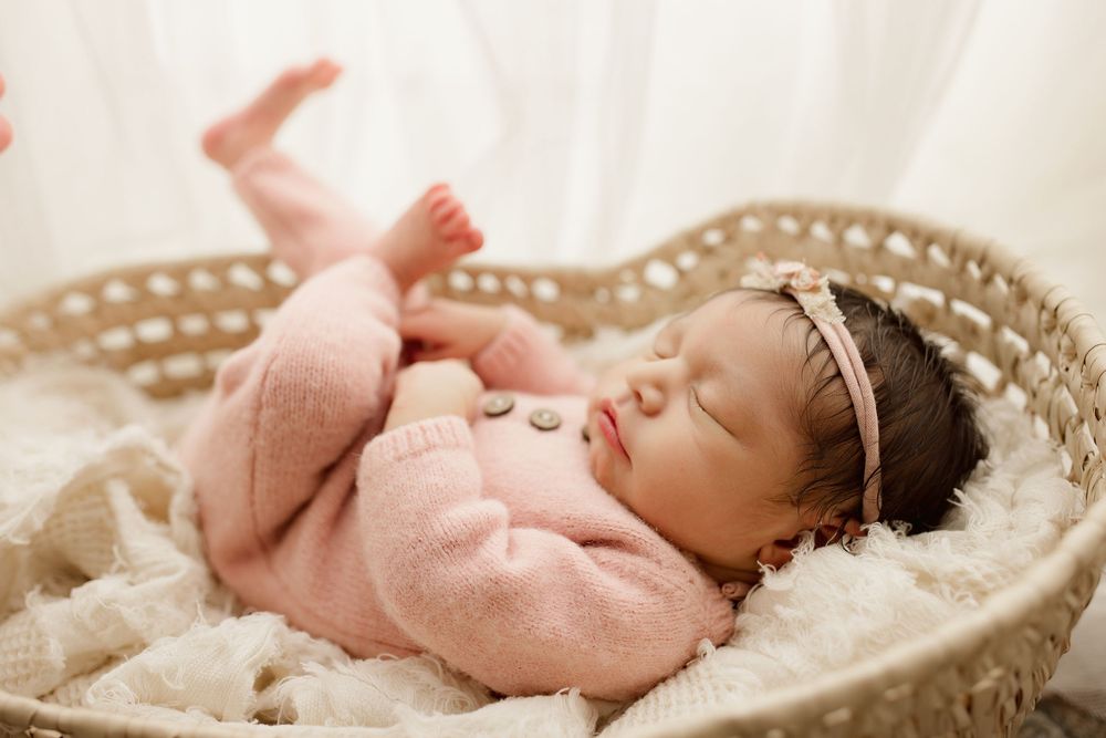 Sleeping newborn baby wearing pink lies peacefully in white woven basket on soft blanket during studio photo session.