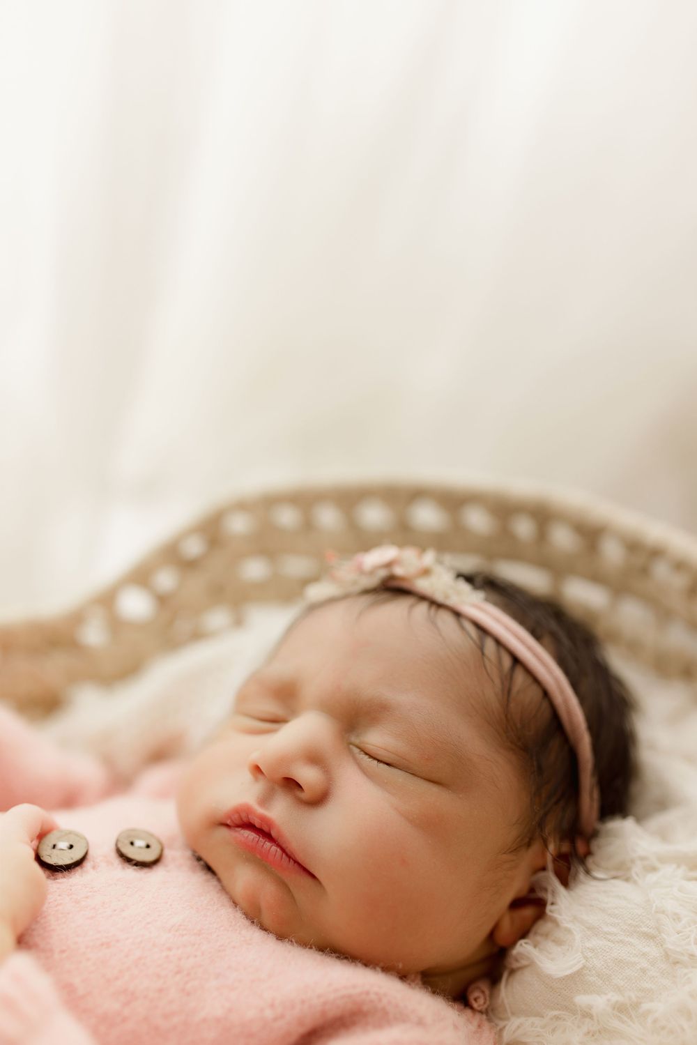 Sleeping newborn wearing pink outfit and headband rests peacefully on soft white blanket.