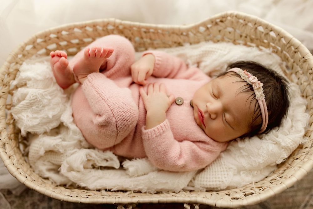 Sleeping newborn wearing soft pink knit outfit rests peacefully in woven basket with cream colored blanket.