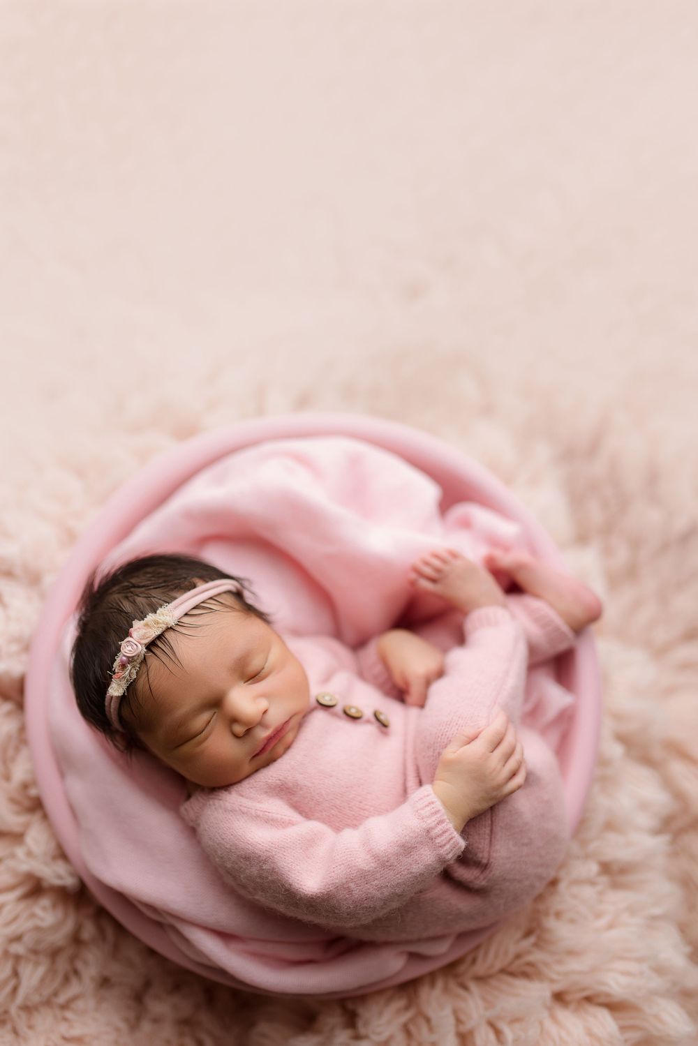 Sleeping newborn wrapped in soft pink blanket nestled in round basket on fluffy beige backdrop.