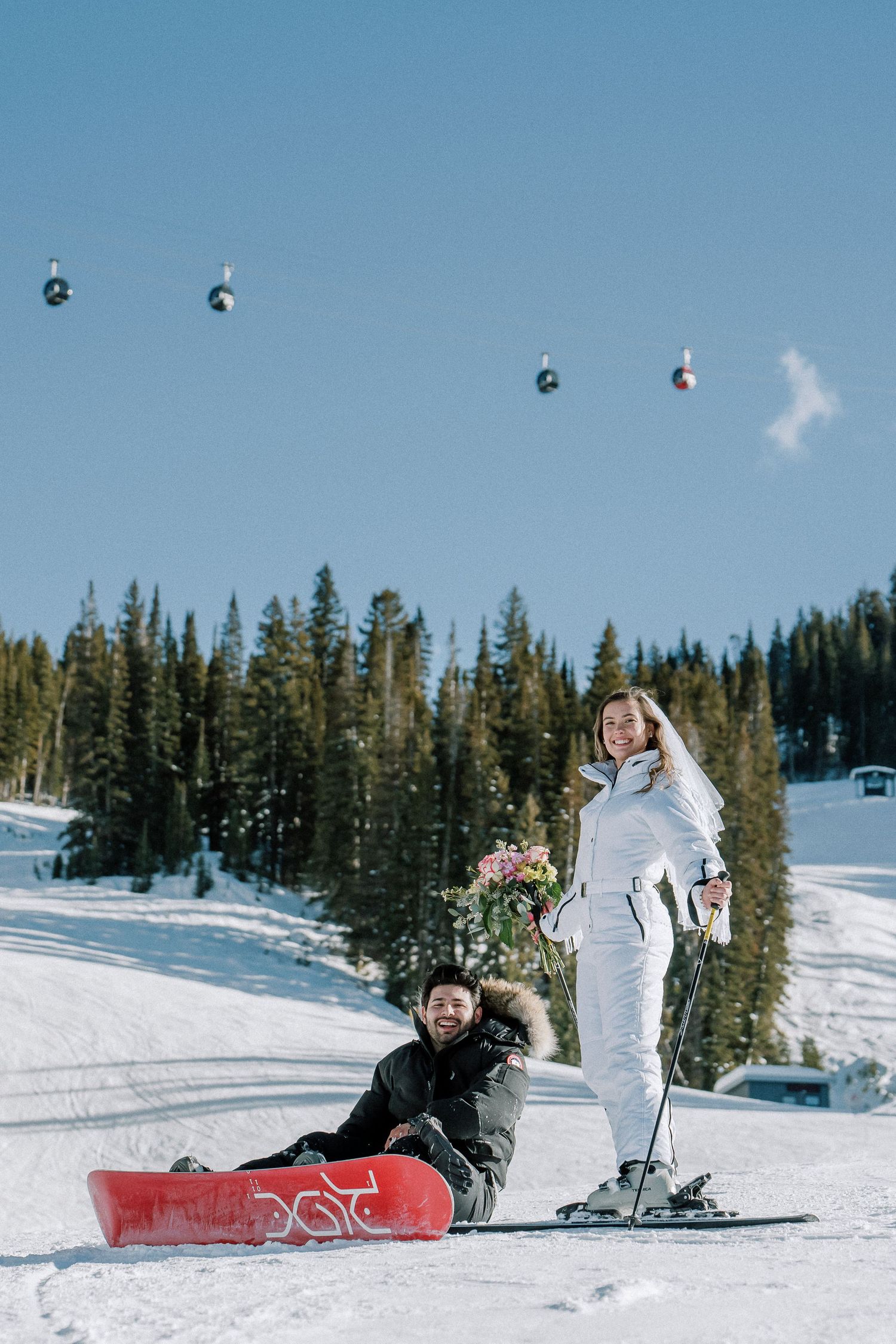 Two people enjoying winter sports activities at a scenic snow covered ski resort with chairlifts and evergreen trees.