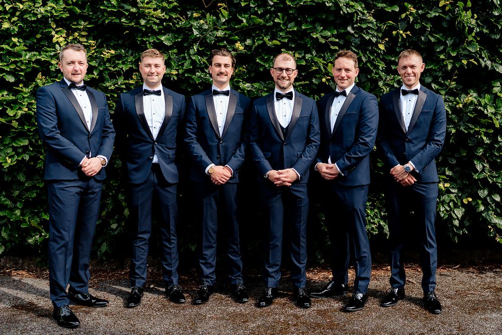 A group of groomsmen in matching navy blue suits and bow ties pose together in front of a leafy green hedge backdrop.