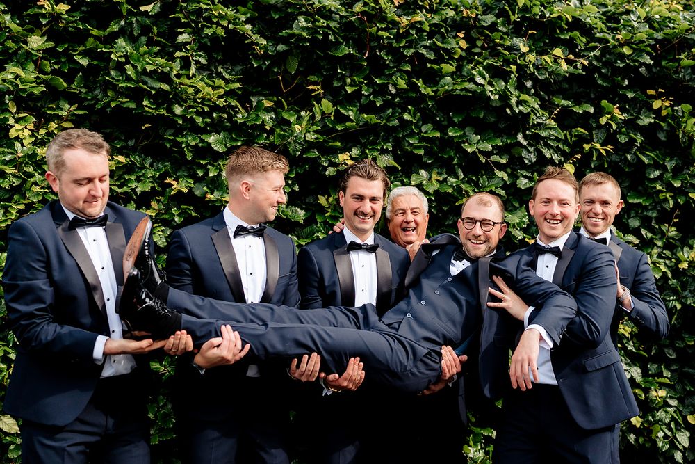 Groomsmen in matching navy suits and bowties playfully carry another groomsman horizontally against a green hedge backdrop.