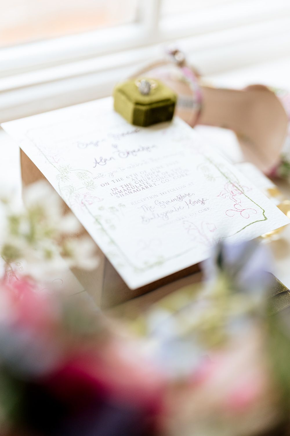 Close-up view of an elegant wedding invitation with soft floral decor and a green heart-shaped box in the background.