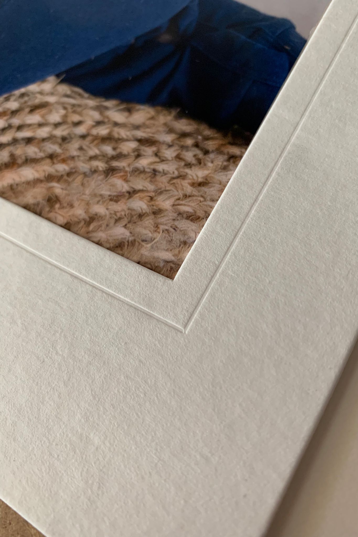 Close-up view of a textured mat board frame corner with partial view of a photograph showing brown knitted fabric.