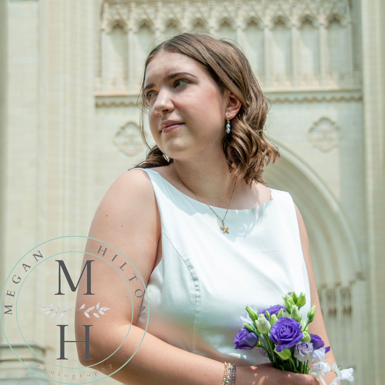 A high school senior in a white sleeveless dress holds a small purple and blue bouquet in front of ornate Gothic church architecture.