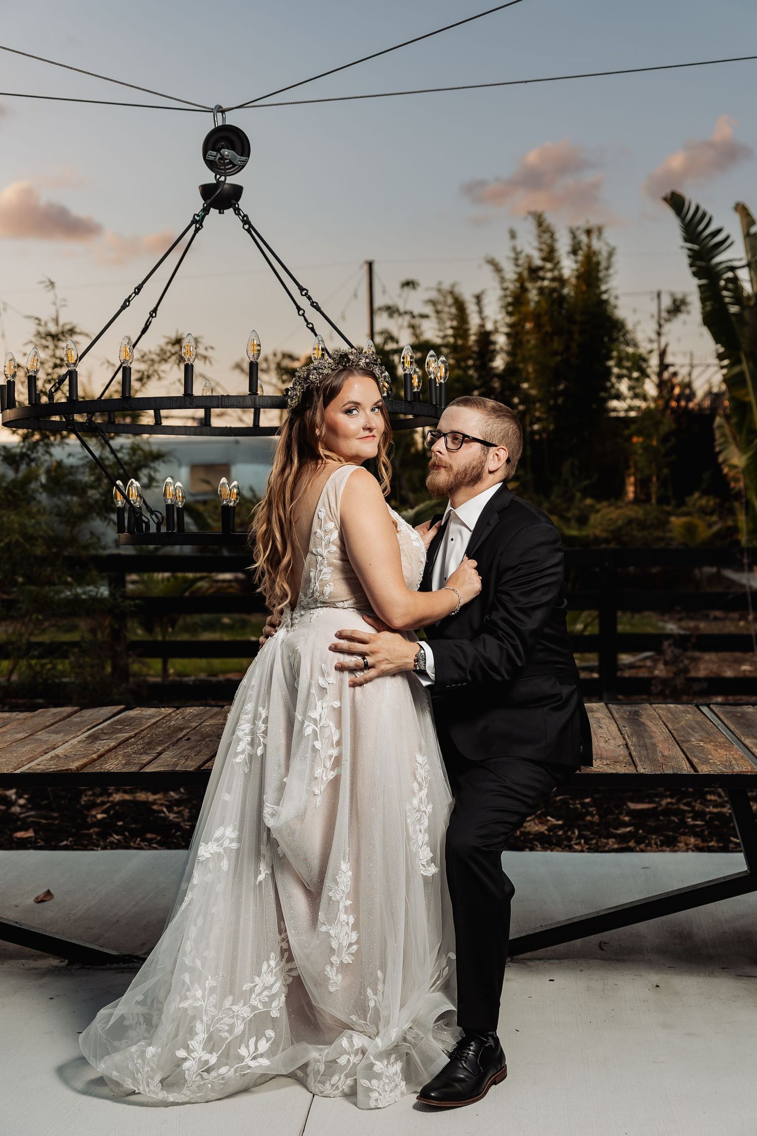 A couple embraces during sunset at an outdoor venue with rustic decor and a vintage industrial chandelier overhead.