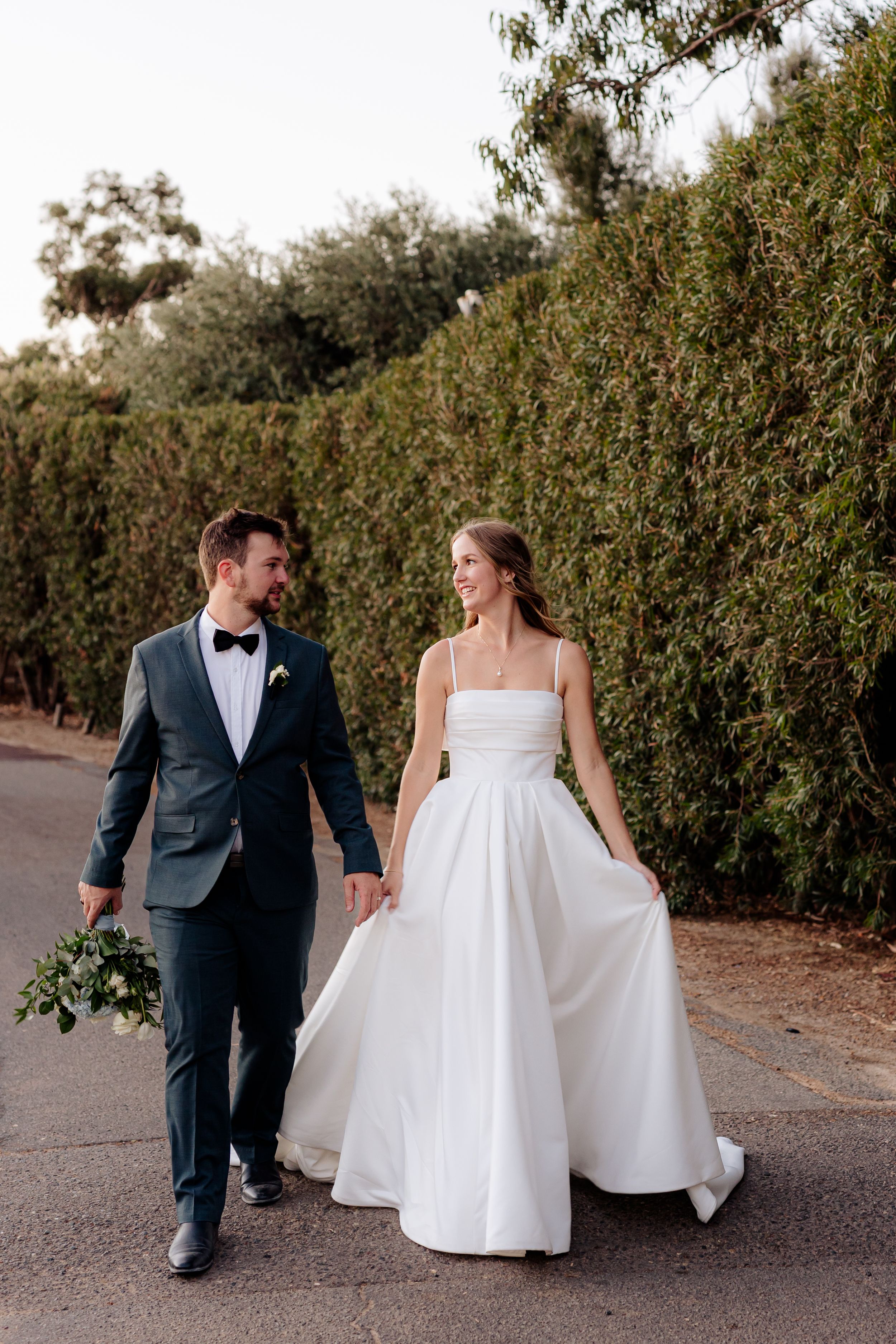A couple on their wedding day walking down a tree-lined path, with the bride in white and groom in charcoal suit.