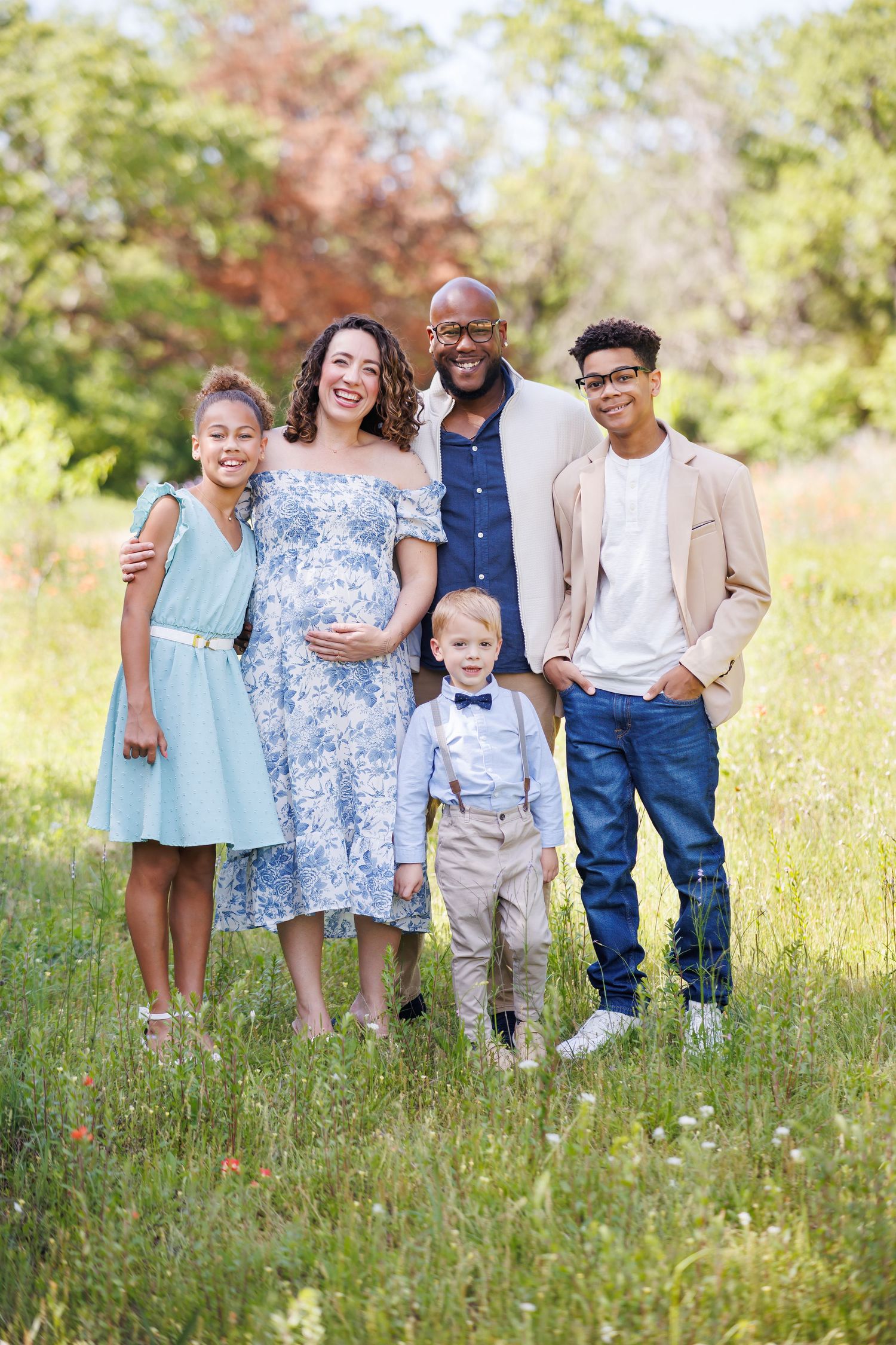 Family of five wearing coordinated blue and neutral tones pose together outdoors in a sunny meadow during spring in Dallas