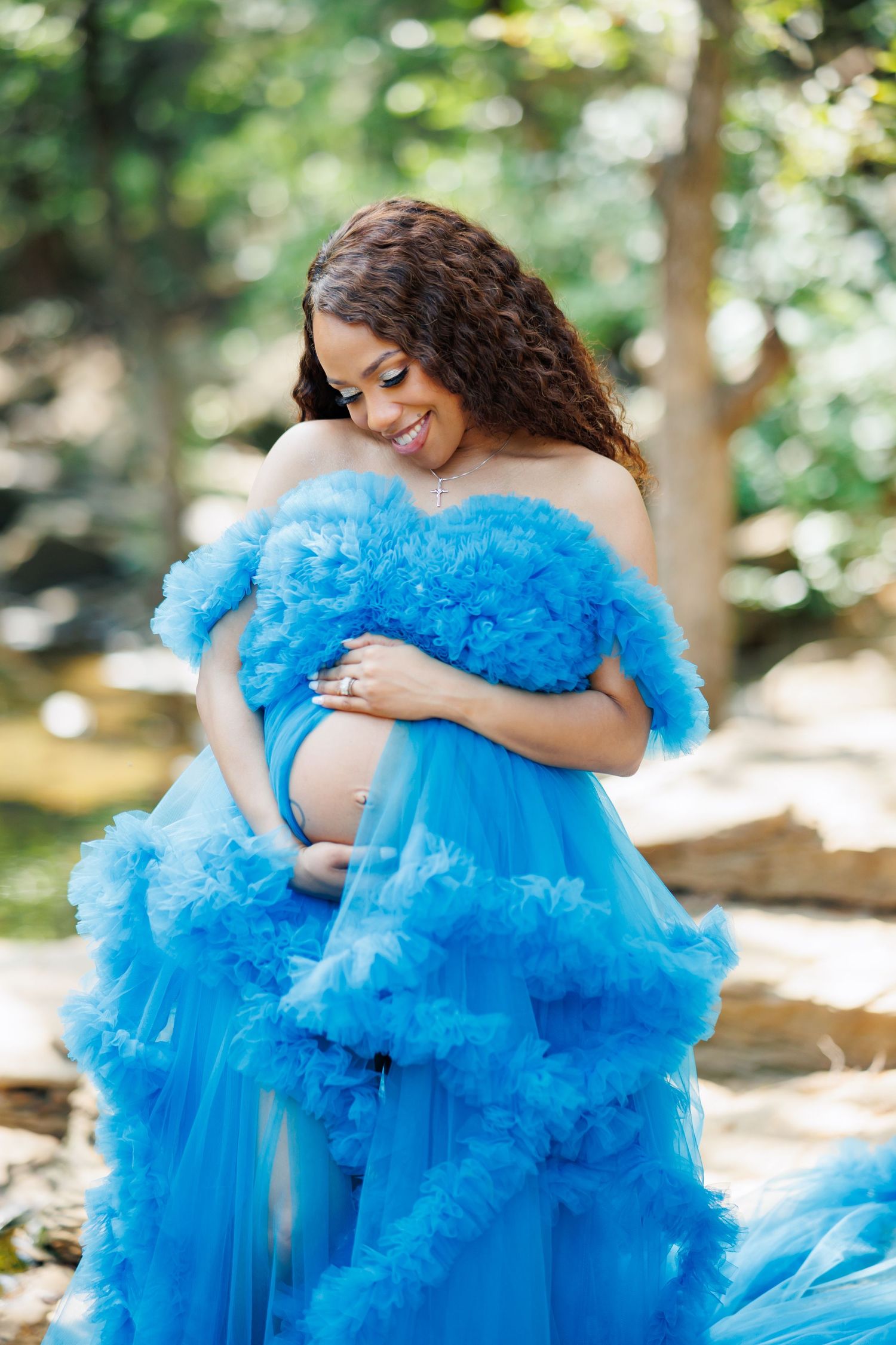 A woman in a flowing blue tulle gown cradles her pregnant belly while standing in a sunlit forest setting for maternity photos in Dallas