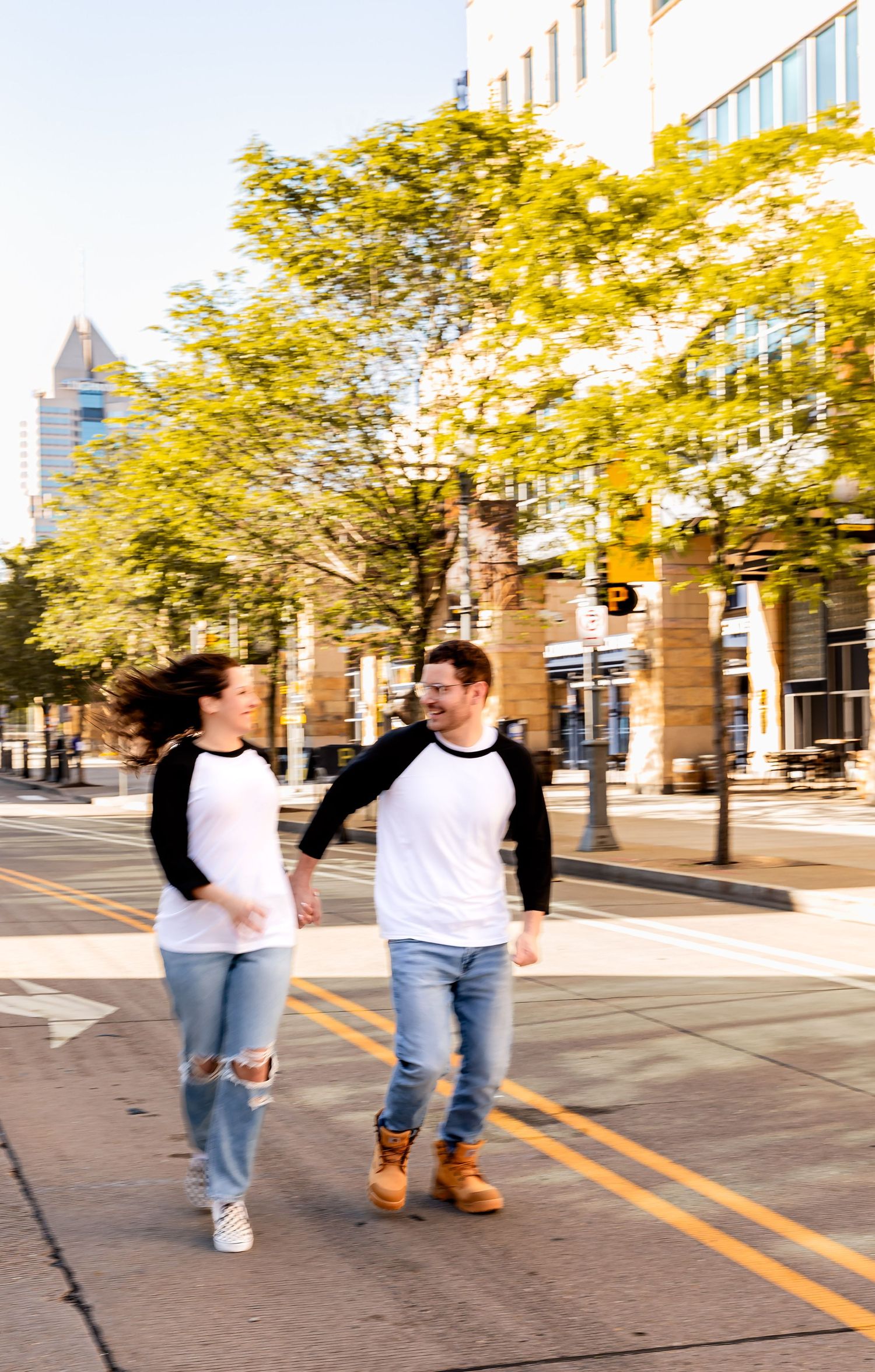 Two people in matching raglan shirts and jeans walk together on a city sidewalk lined with autumn trees.