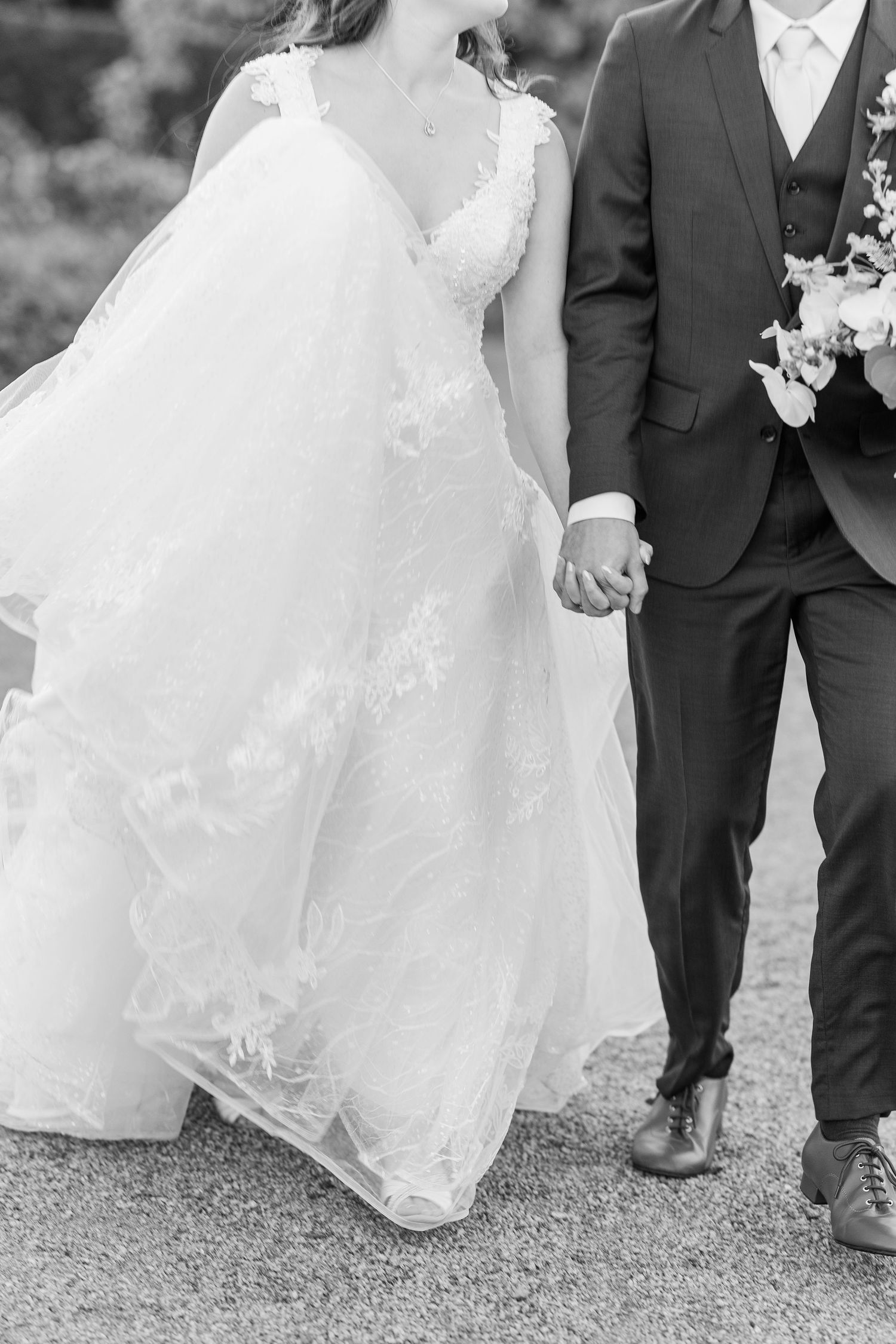A black and white detail shot of a bride in a lace wedding dress holding hands with a groom in a dark suit.