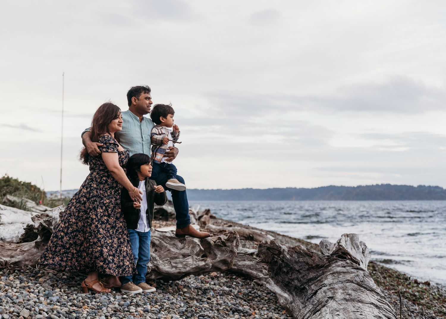 A family enjoys a cloudy day together on a rocky beach with mountains visible across the water.