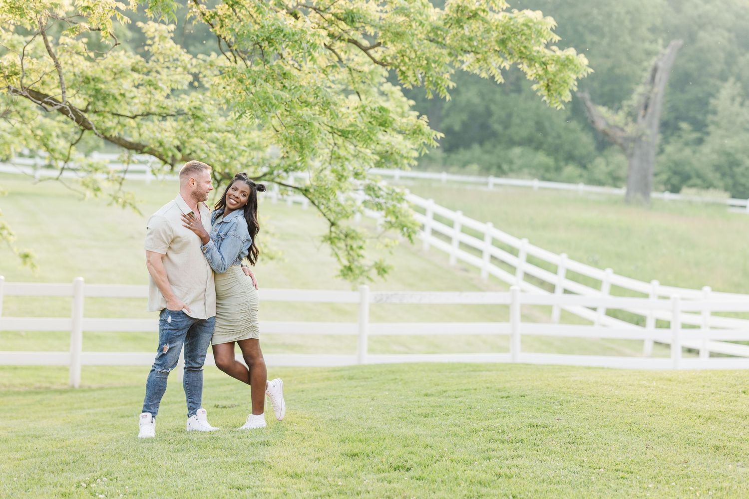 A couple stands together against white fencing in a lush green field during a sunny afternoon.