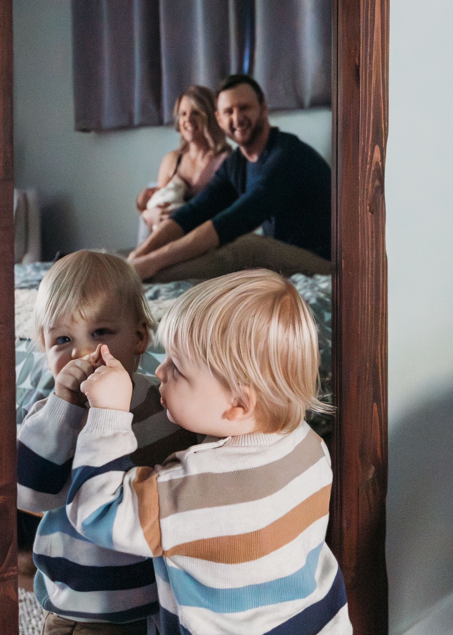 Two young children in striped sweaters play in foreground while their parents sit together on bed in background.