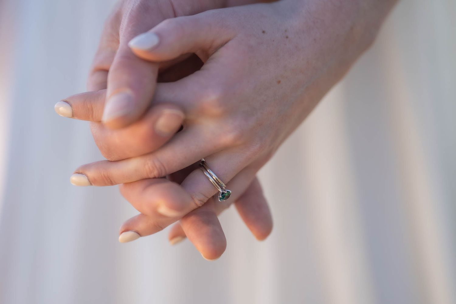 Delicate hands with manicured nails displaying an elegant engagement ring against a light background.