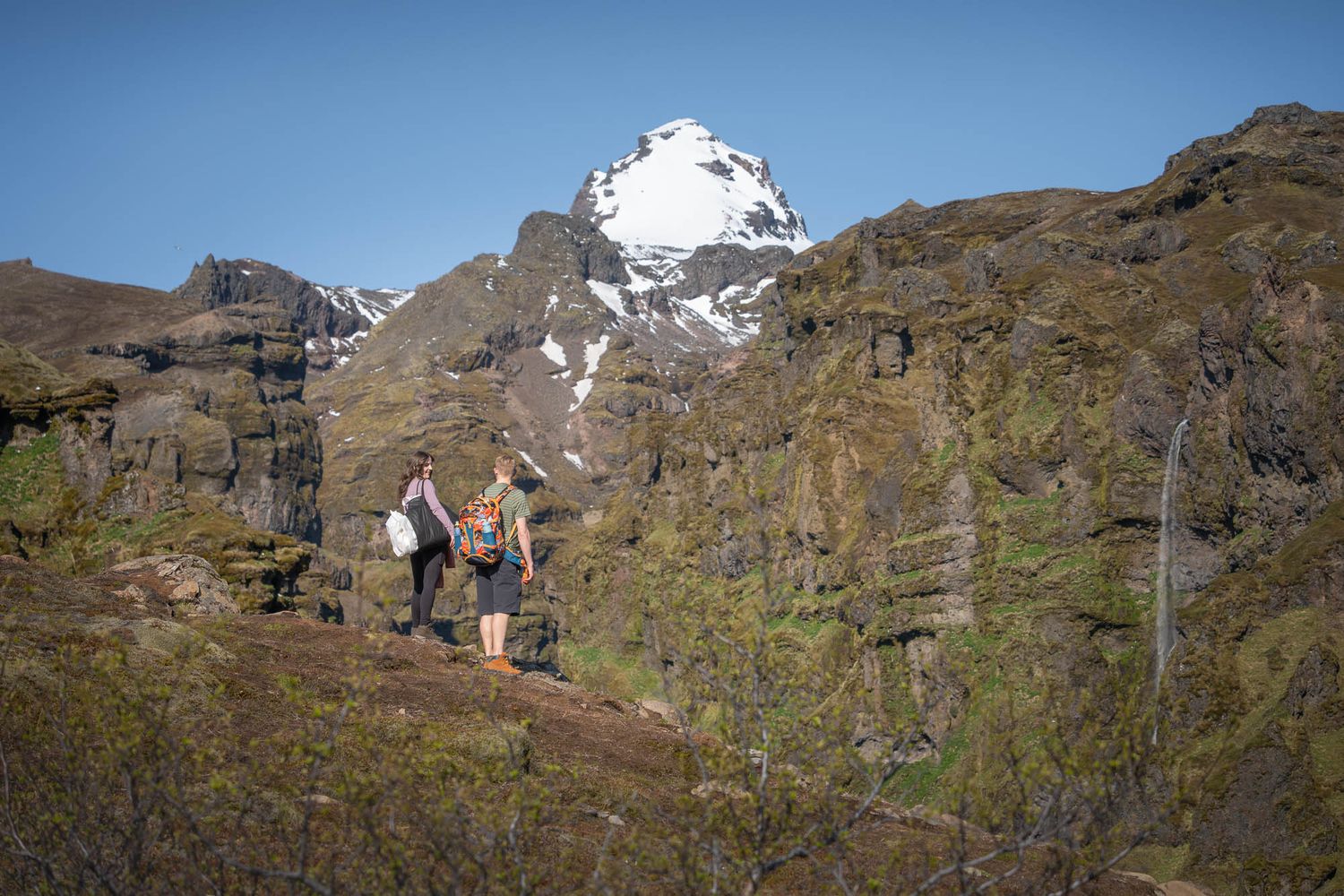 A couple hikes on a mountain trail with snow-capped peaks in the background.