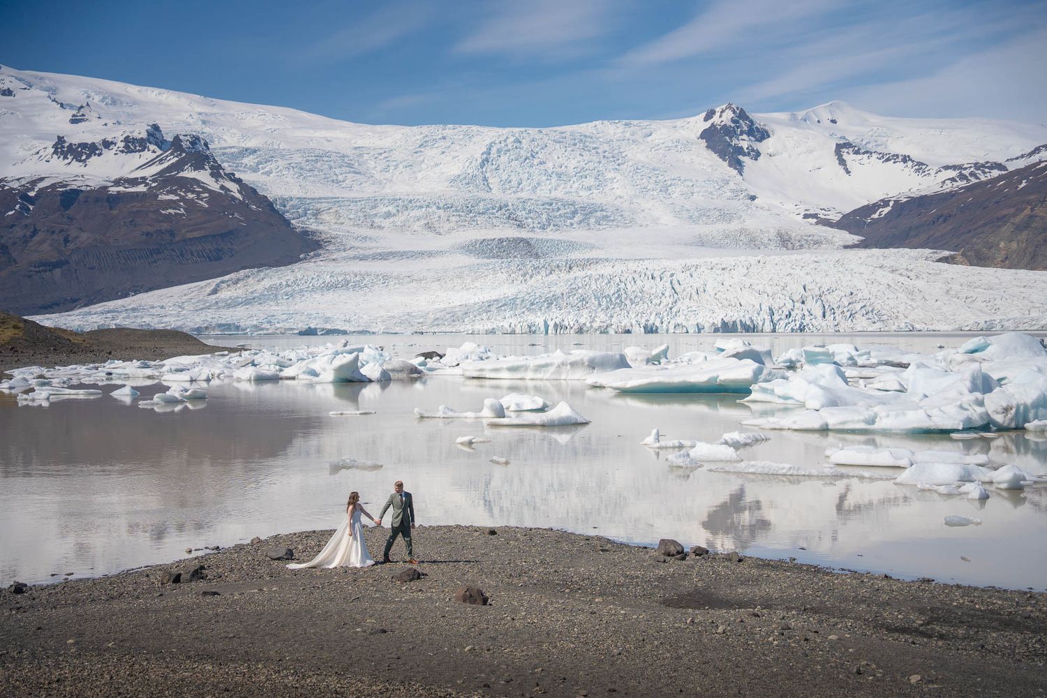 Couple standing on shore with dramatic glacier and icebergs in background.