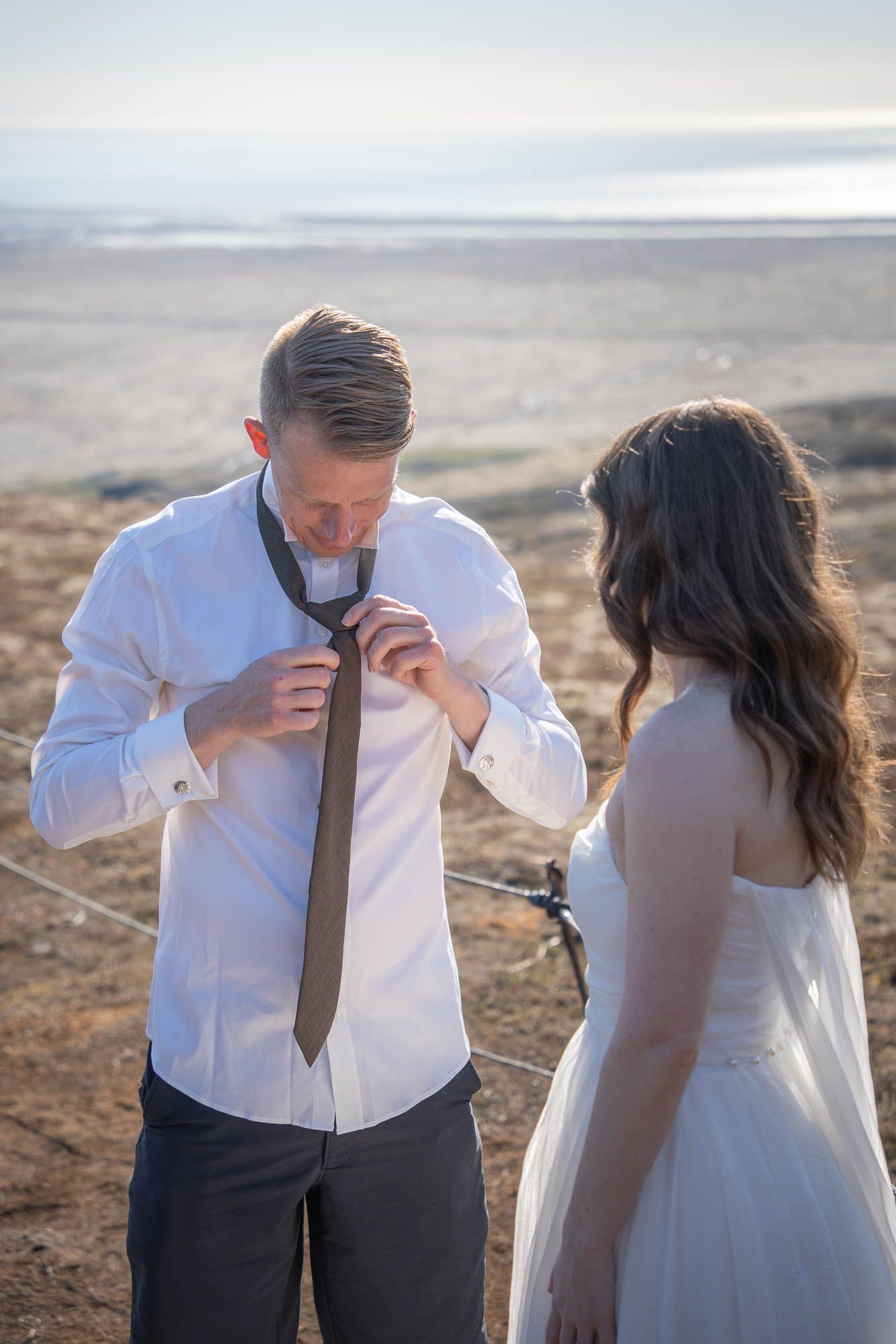 Two people in wedding attire stand in a desert landscape as the groom adjusts his tie at sunset.