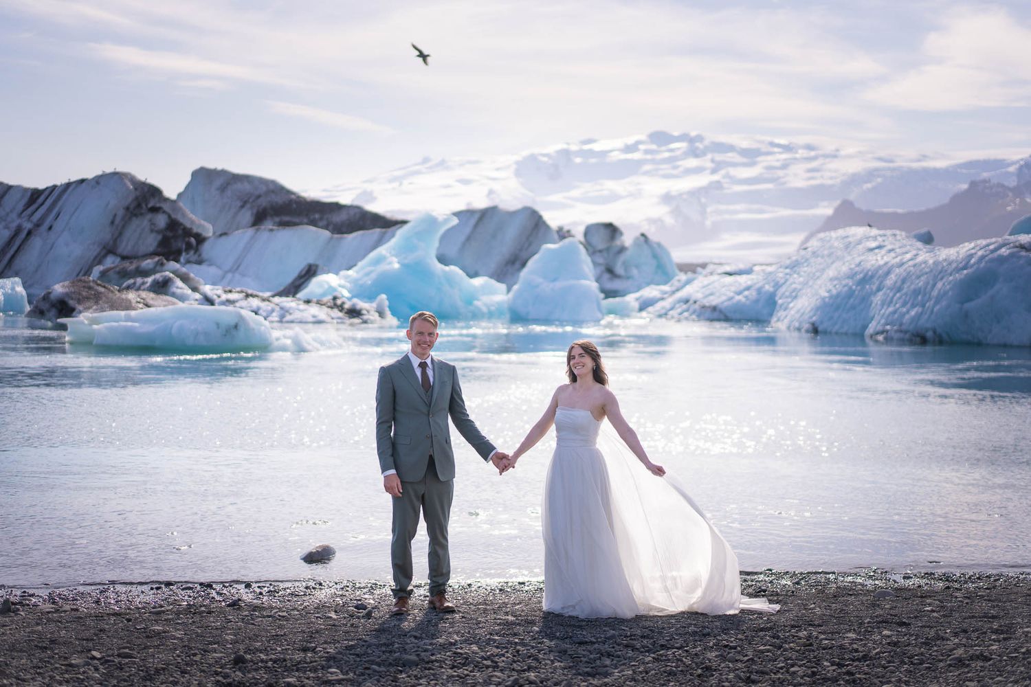 Couple in wedding attire holding hands in front of stunning blue icebergs at a glacial lagoon.