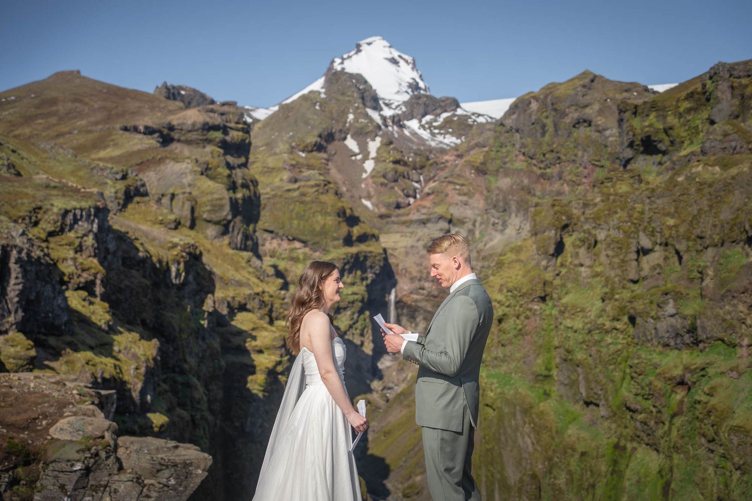 Wedding couple standing on dramatic mountain cliffs with snow-capped peaks in the scenic backdrop.