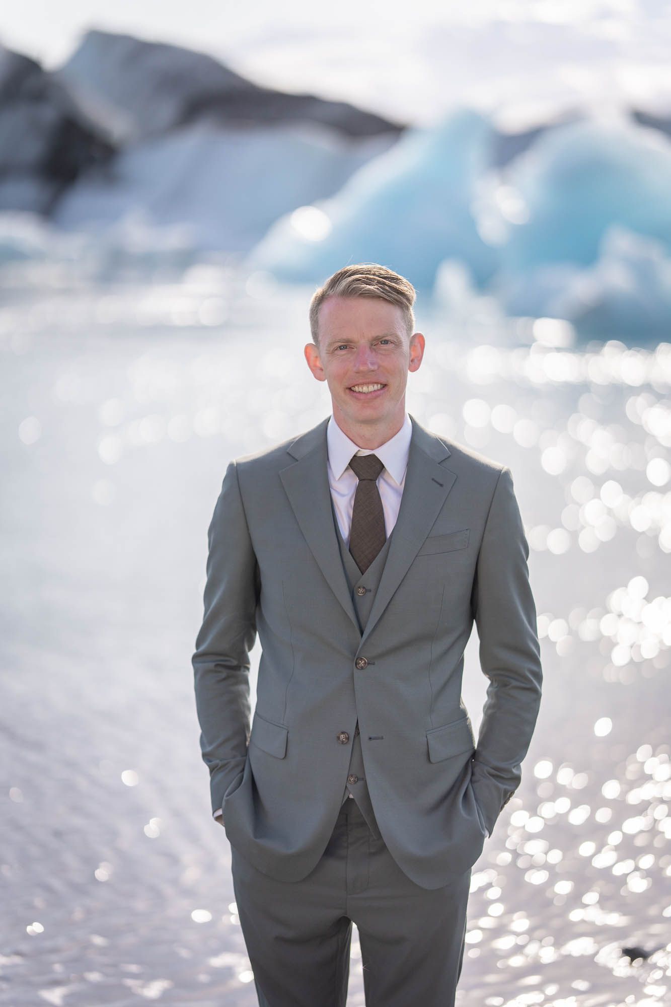 Well-dressed person in a gray suit poses against a stunning glacial backdrop with sparkling water.