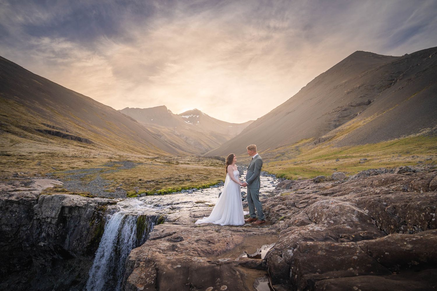 A wedding couple photographed in front of a dramatic mountain valley with waterfalls at sunset.