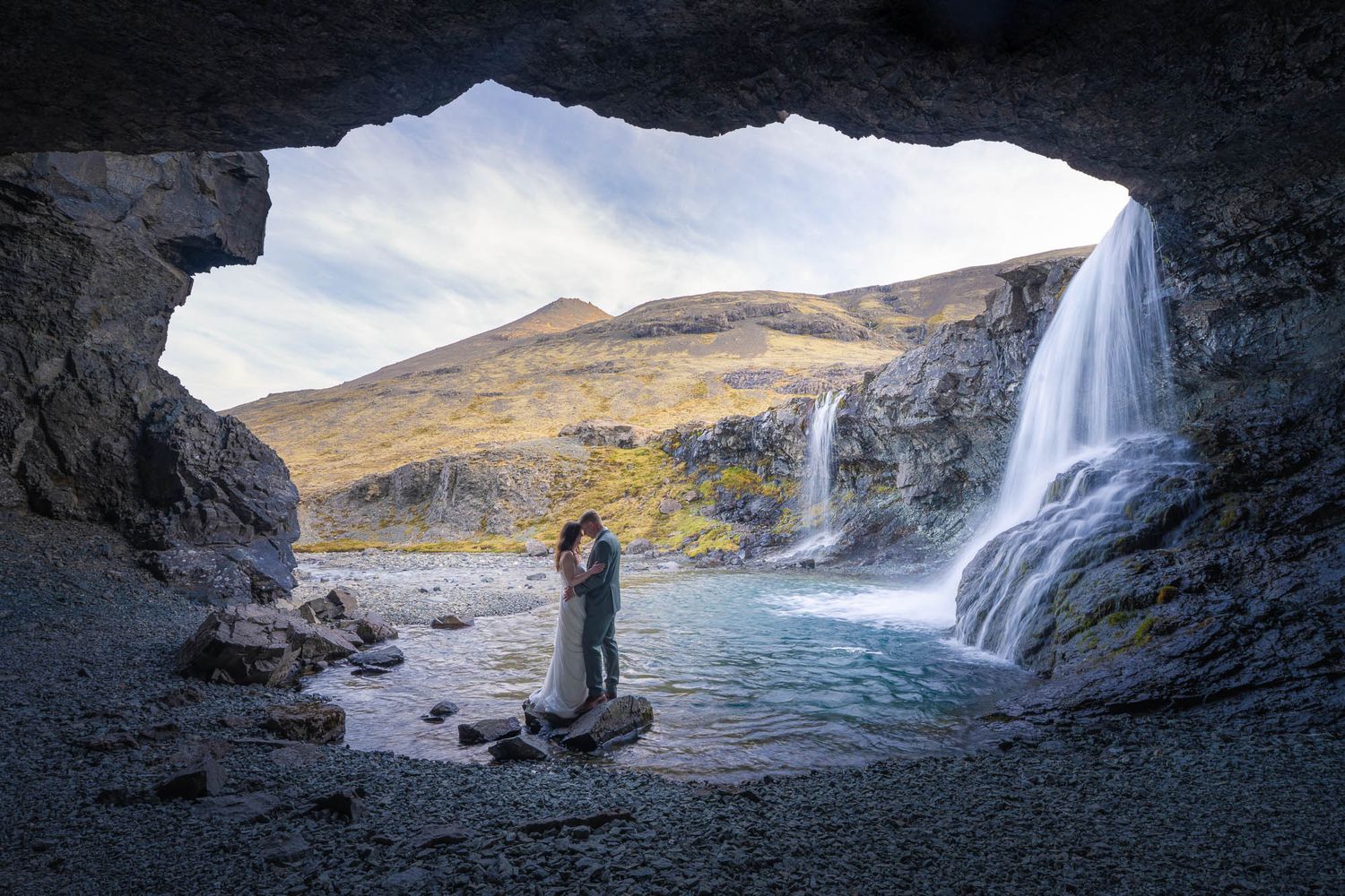 Silhouette of embracing couple inside a cave opening with waterfall and mountains visible beyond.