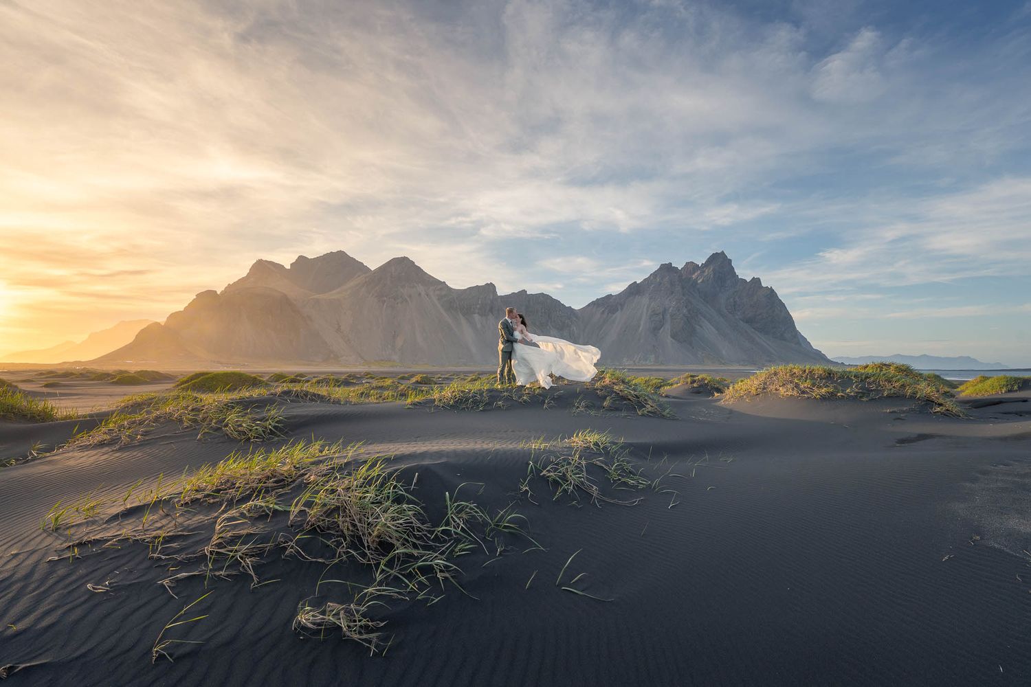 A silhouetted couple stands on dark sand dunes against majestic mountain peaks during a golden sunset in Iceland.