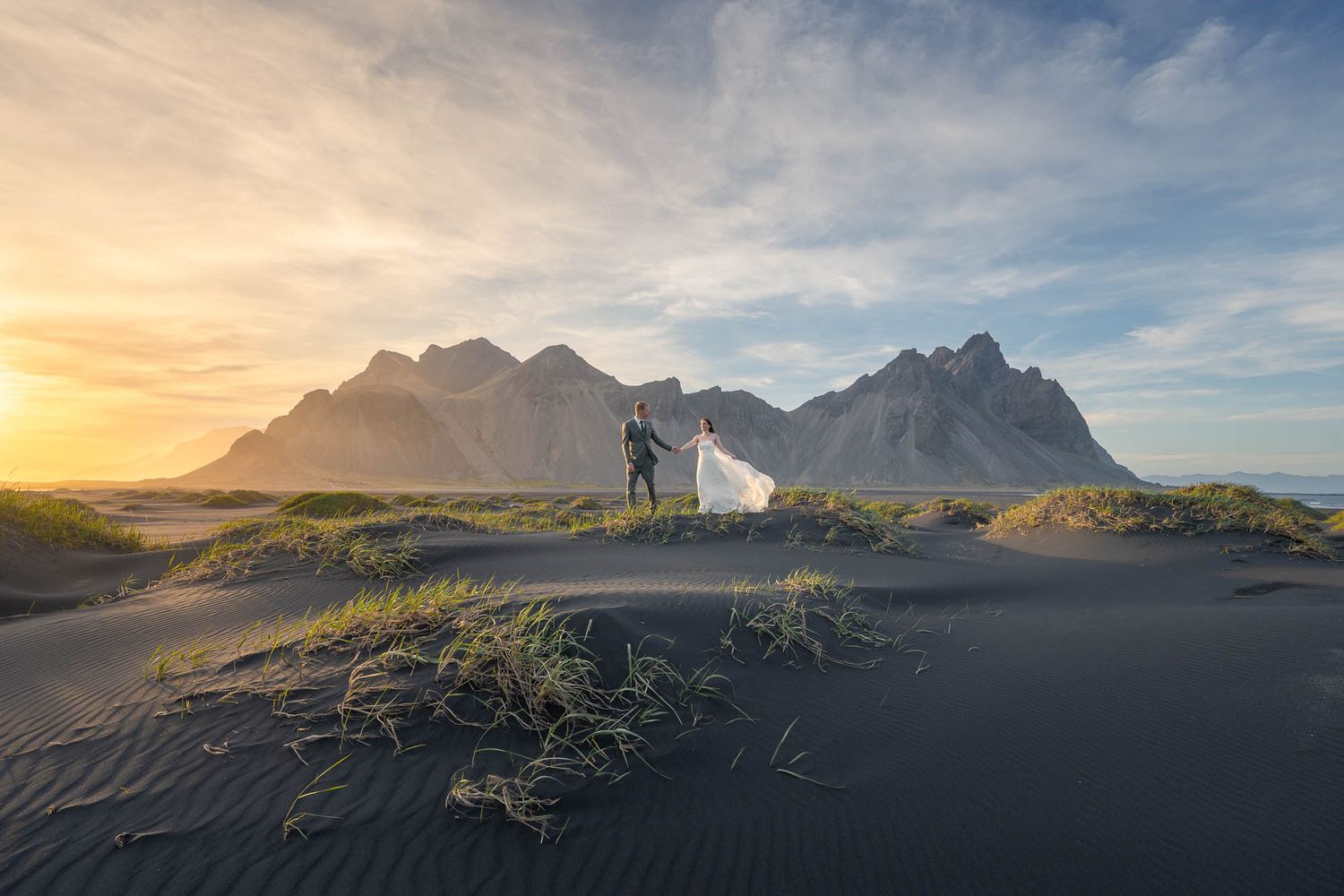 Dramatic sunset view of Vestrahorn mountains rising above black sand dunes in Iceland with golden light and scattered clouds.