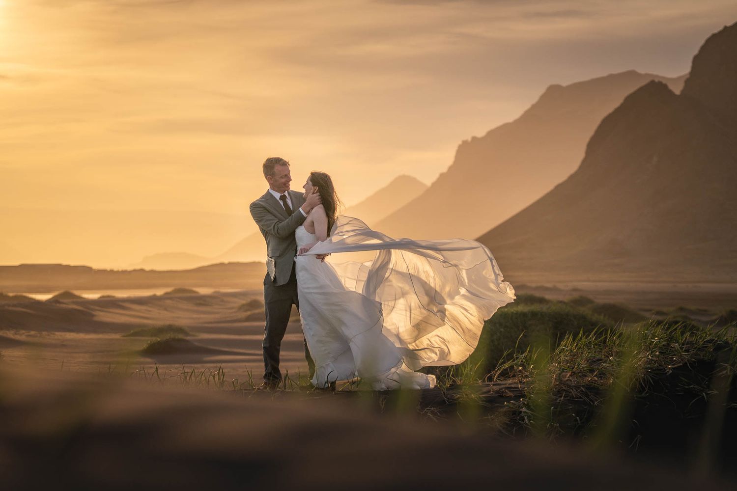 A couple in wedding attire poses dramatically against a golden sunset with mountains and billowing veil in the background.