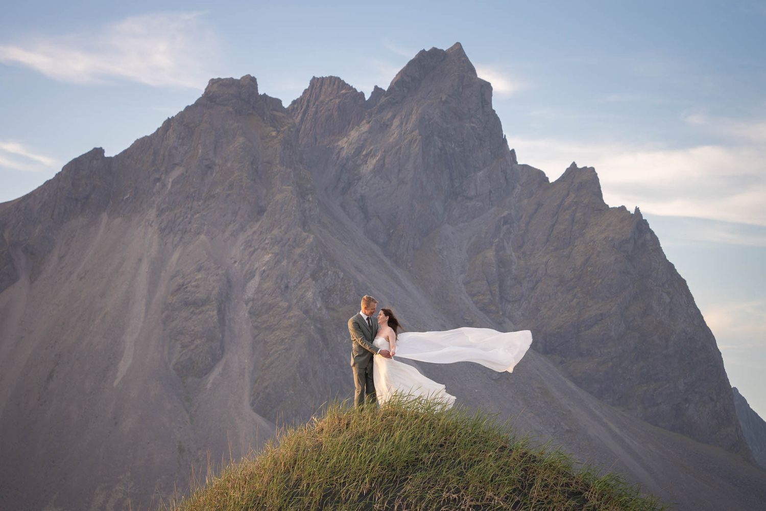 A couple embraces on a hilltop against dramatic mountain peaks as the bride's white dress flows in the wind.