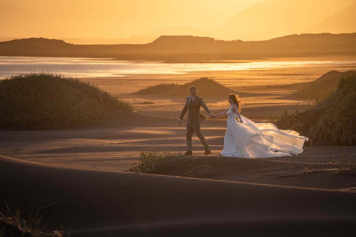 Two people in wedding attire hold hands while walking across sand dunes during a golden sunset.