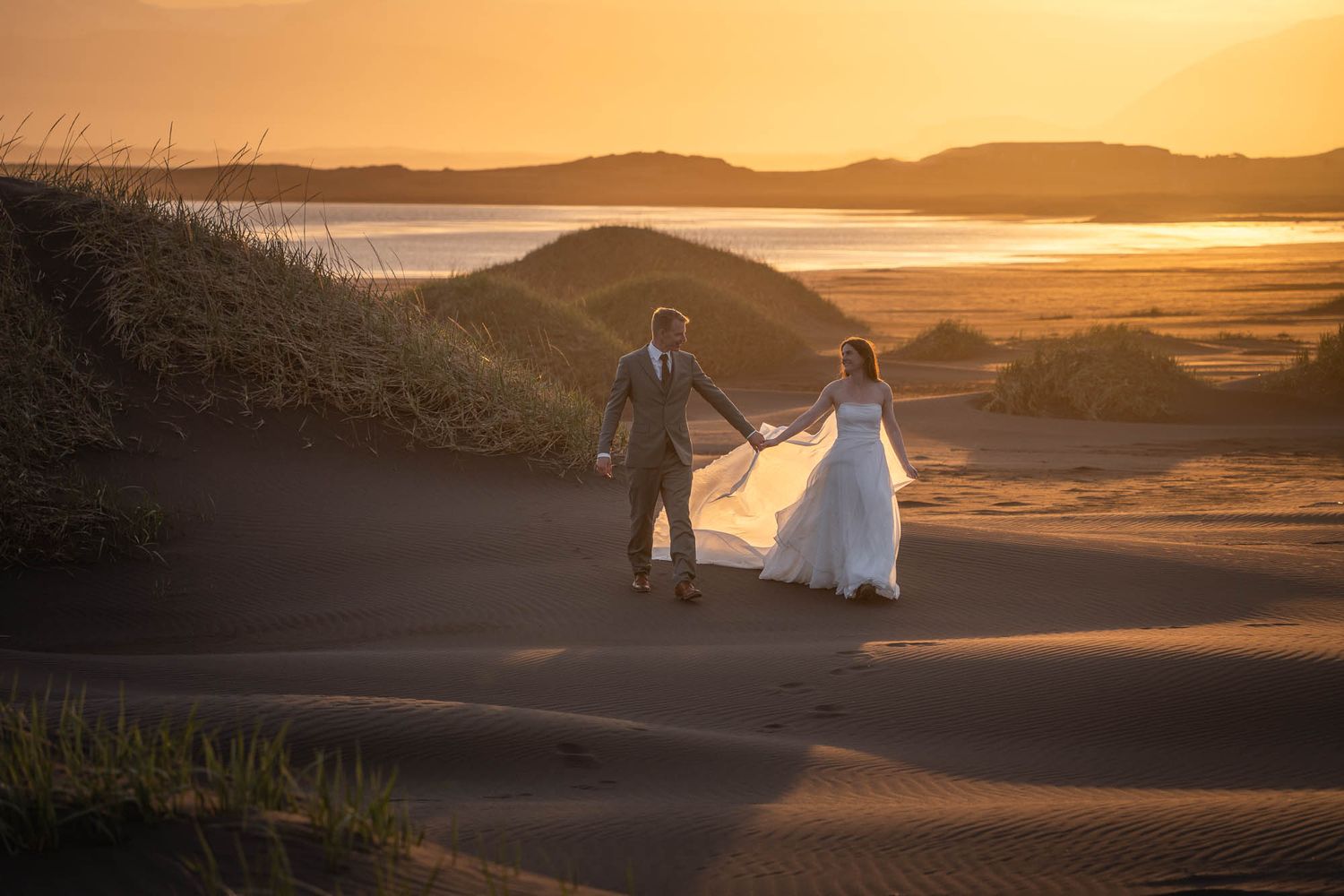 A couple walks along coastal sand dunes at sunset with glowing warm light and ocean views.