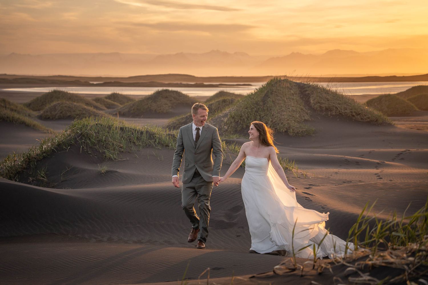 Couple walking hand in hand through sand dunes during golden hour sunset.