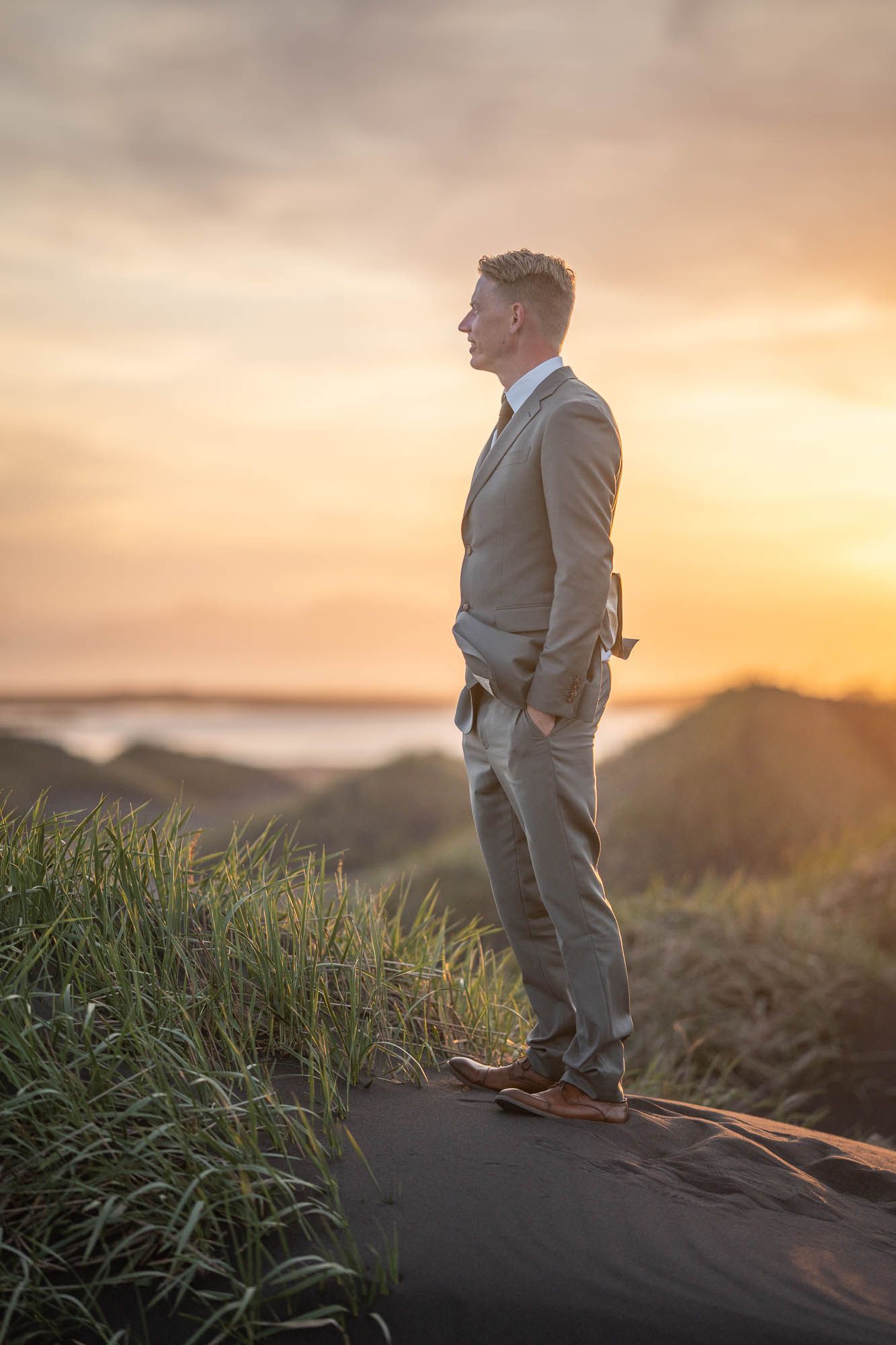 A person in a light suit stands on a cliff edge looking out at a dramatic coastal sunset.