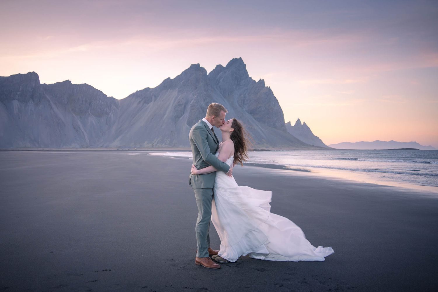 Couple embracing on a black sand beach with dramatic mountain peaks and sunset backdrop in Iceland.
