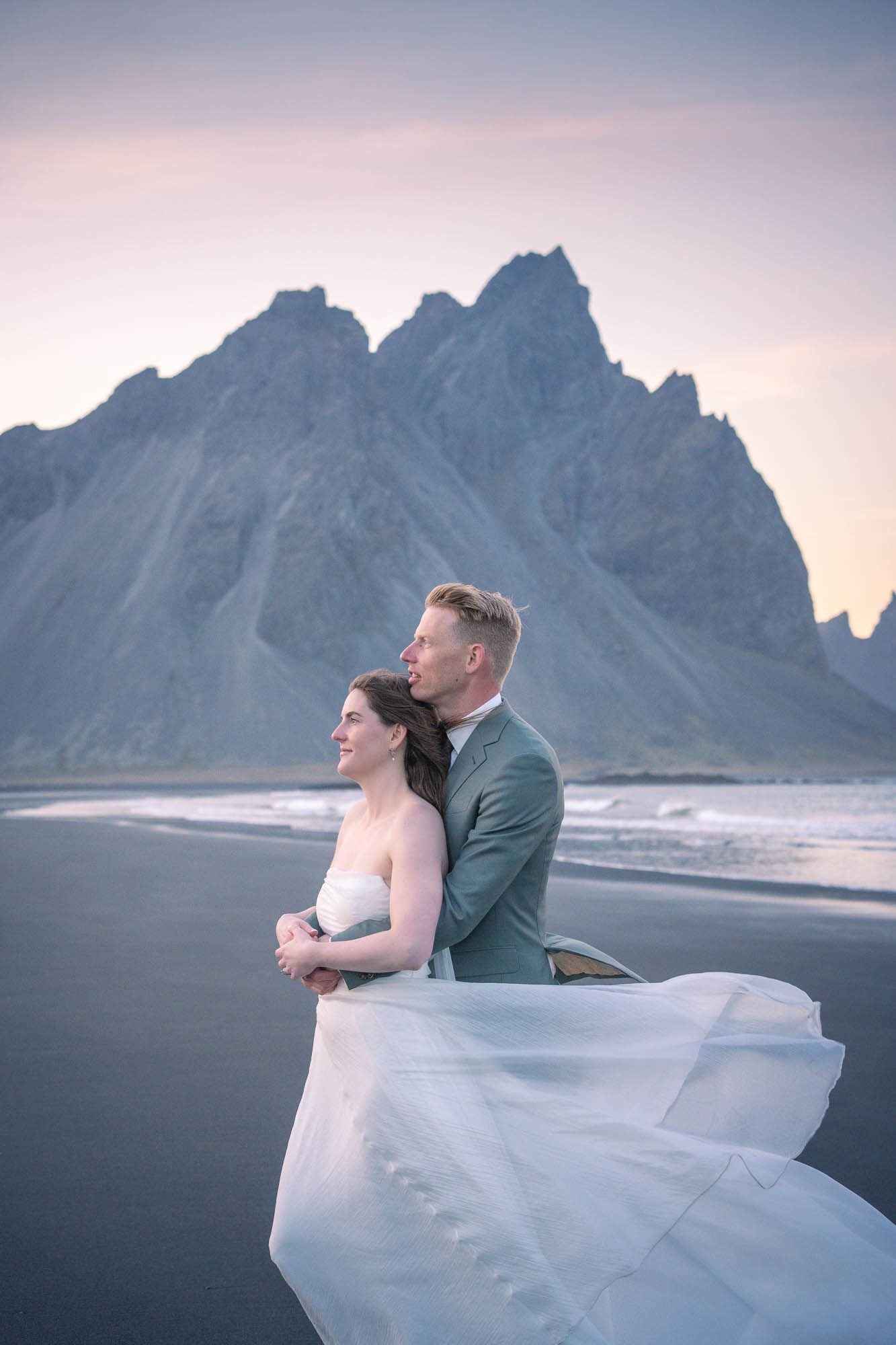 A couple embraces on a black sand beach with dramatic mountain peaks of Vestrahorn in Iceland during sunset.