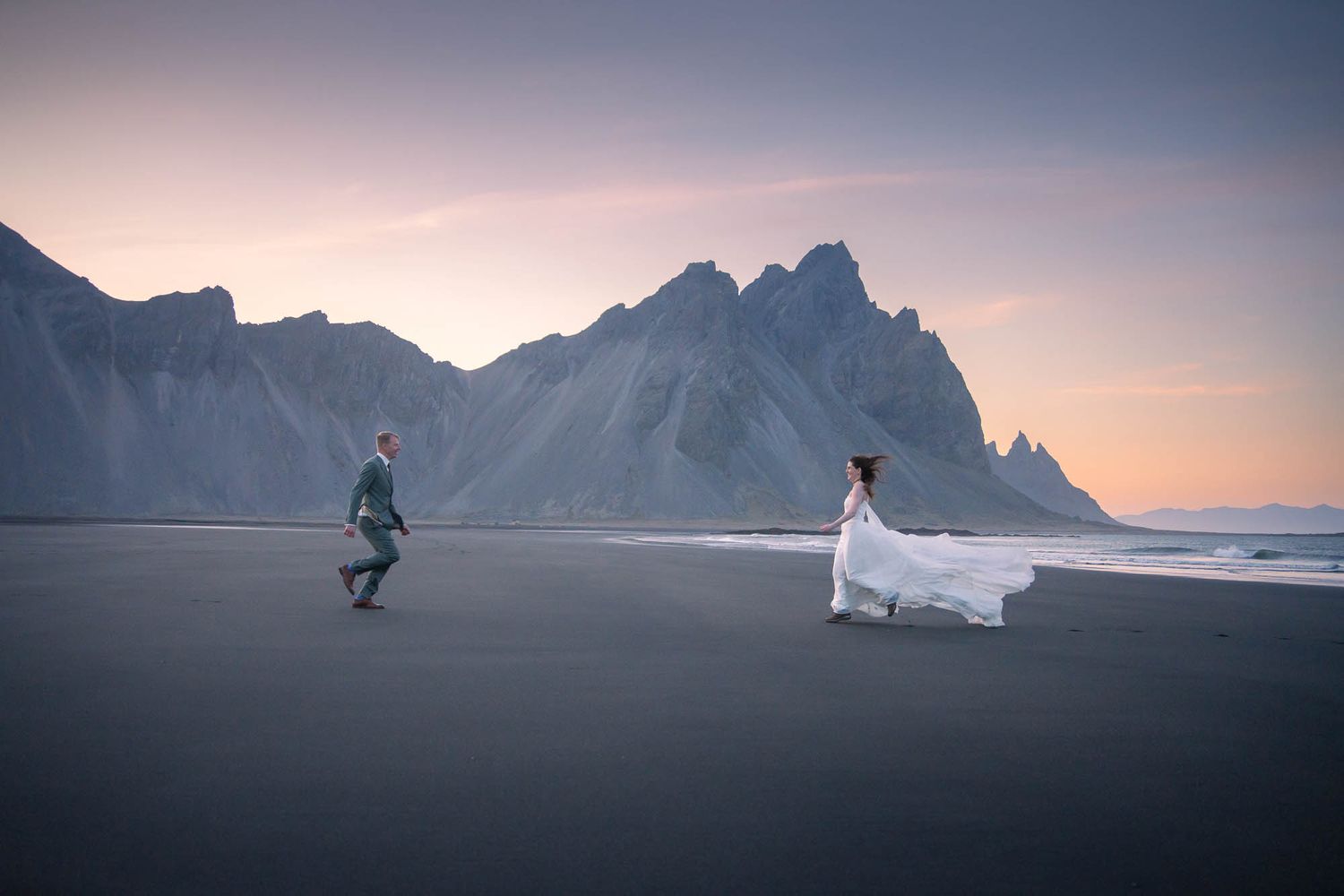 Two figures move across a black sand beach with dramatic mountain peaks and pink sky in the background.