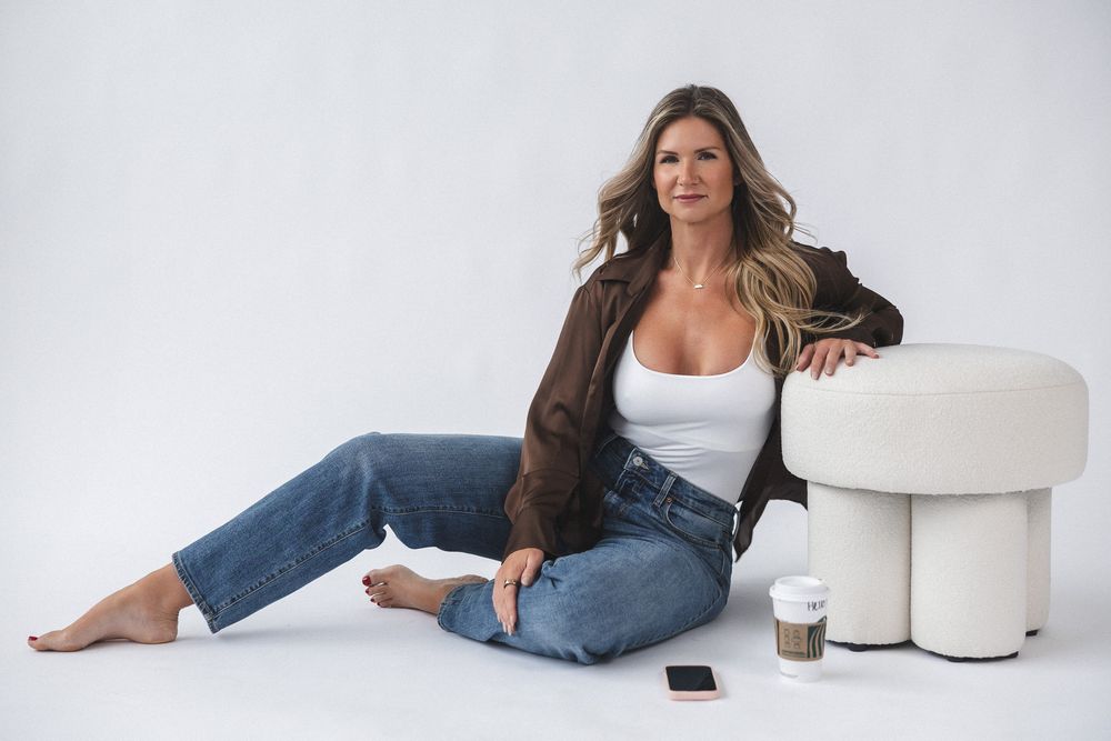 Casual portrait of a model in denim and brown jacket sitting on floor next to white furniture.