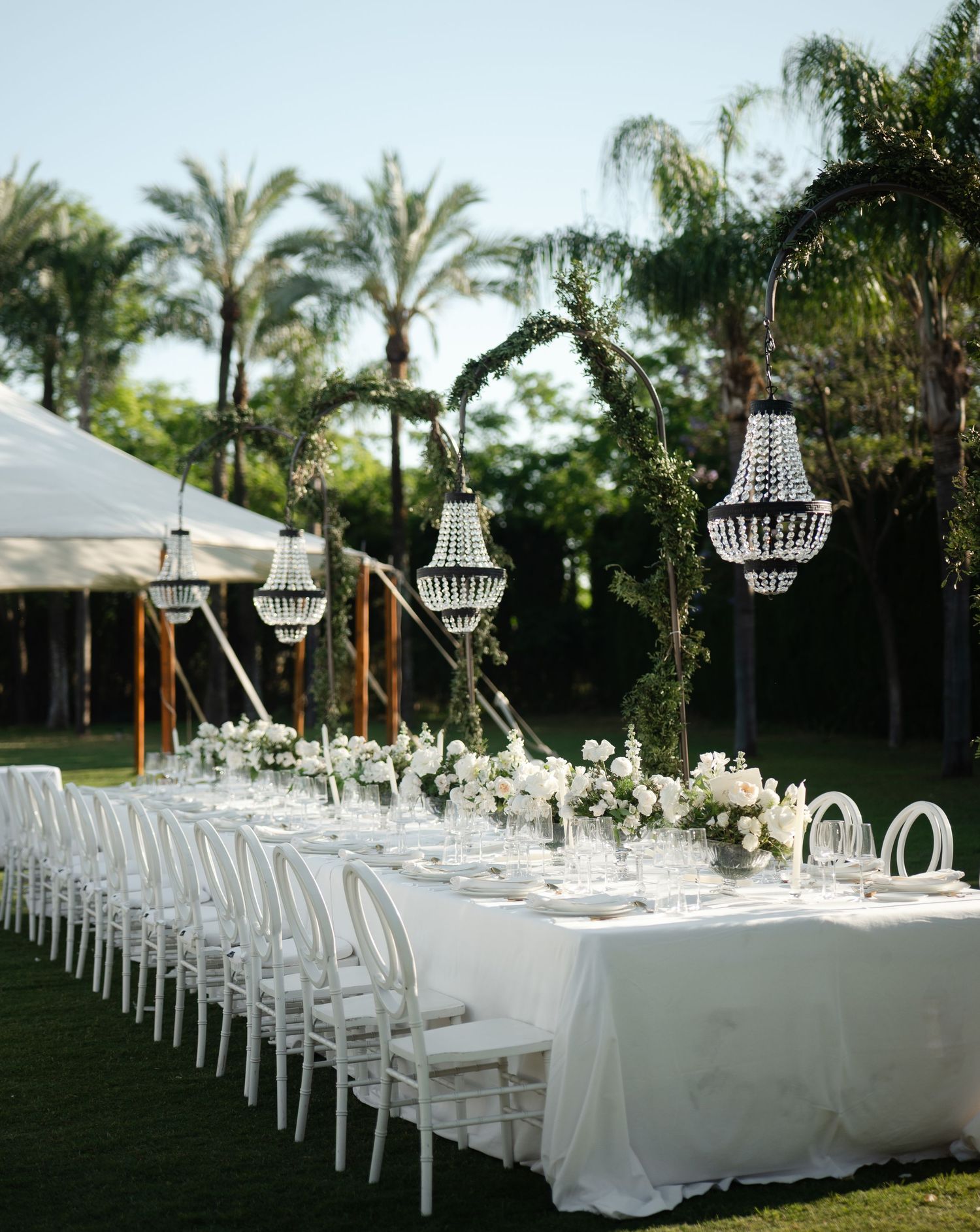 Elegant outdoor wedding table setup with crystal chandeliers hanging from greenery arch and ghost chairs under palm trees.
