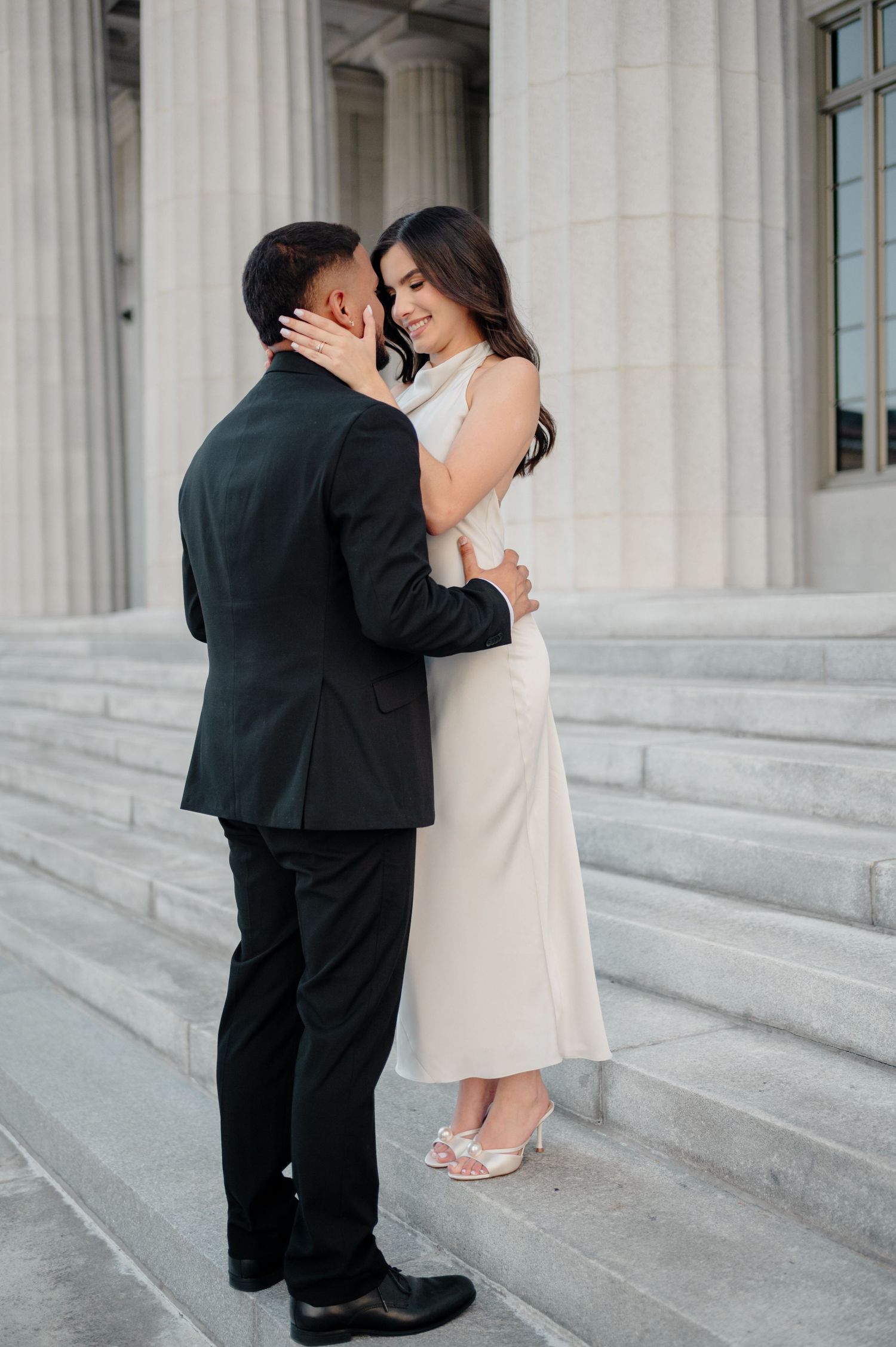 A romantic couple embraces on marble steps in formal attire, with classical columns in the background.