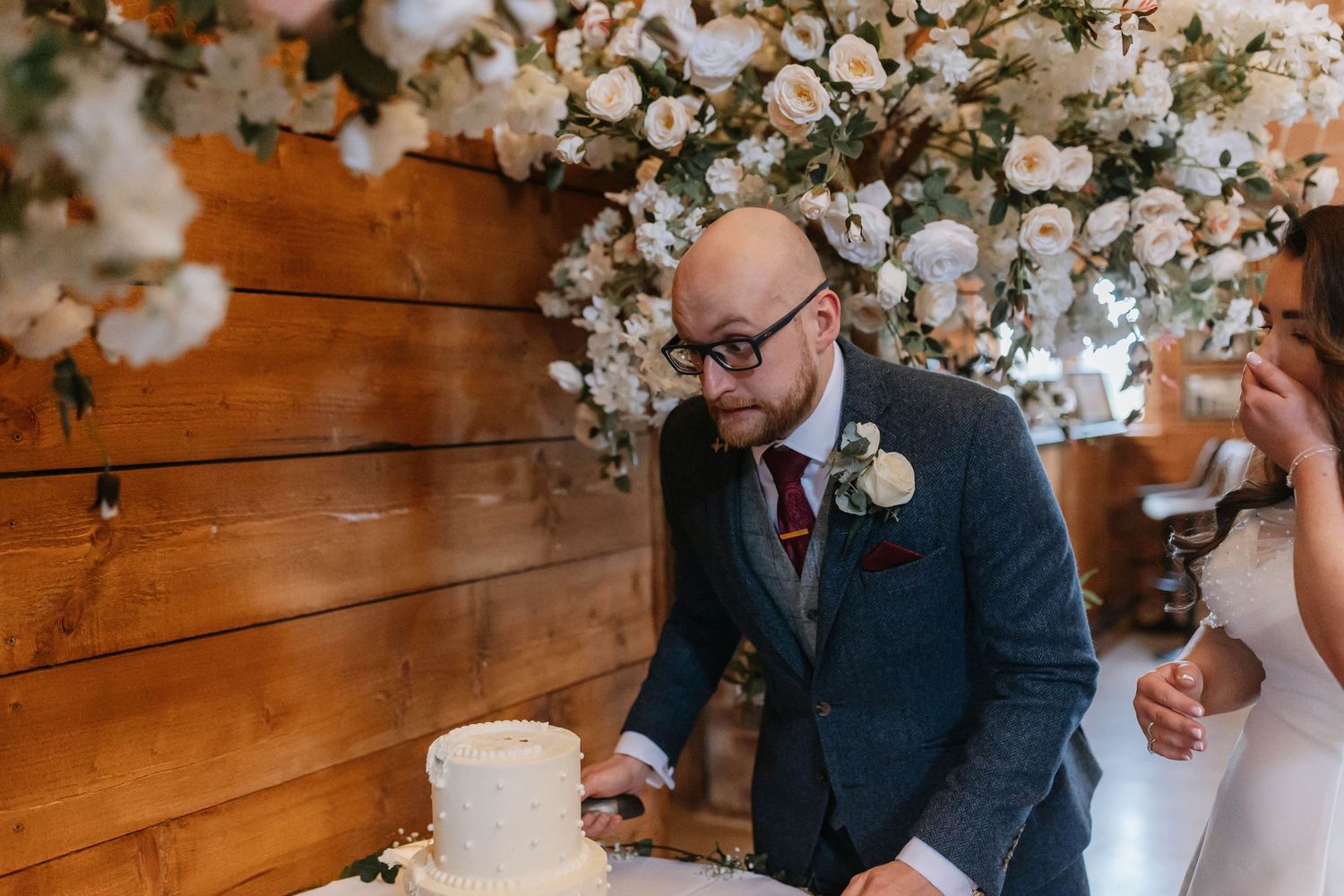 A couple cuts their white wedding cake beneath a beautiful white floral arch against a rustic wooden wall backdrop.