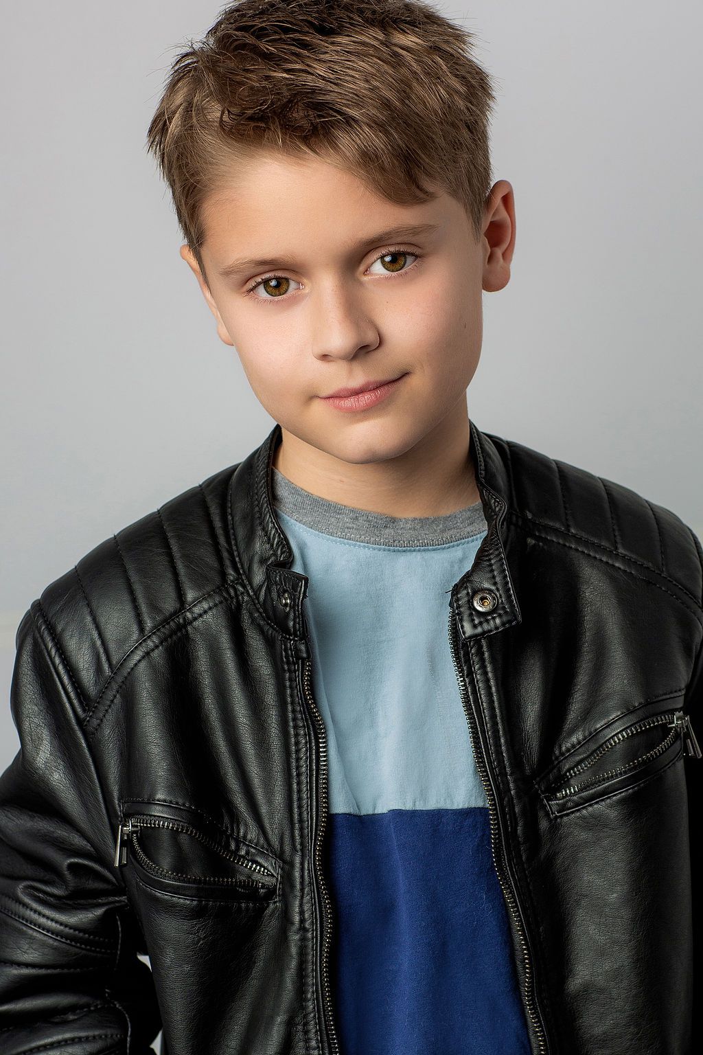 A young person in a black leather jacket and blue shirt poses against a gray background for a headshot portrait.