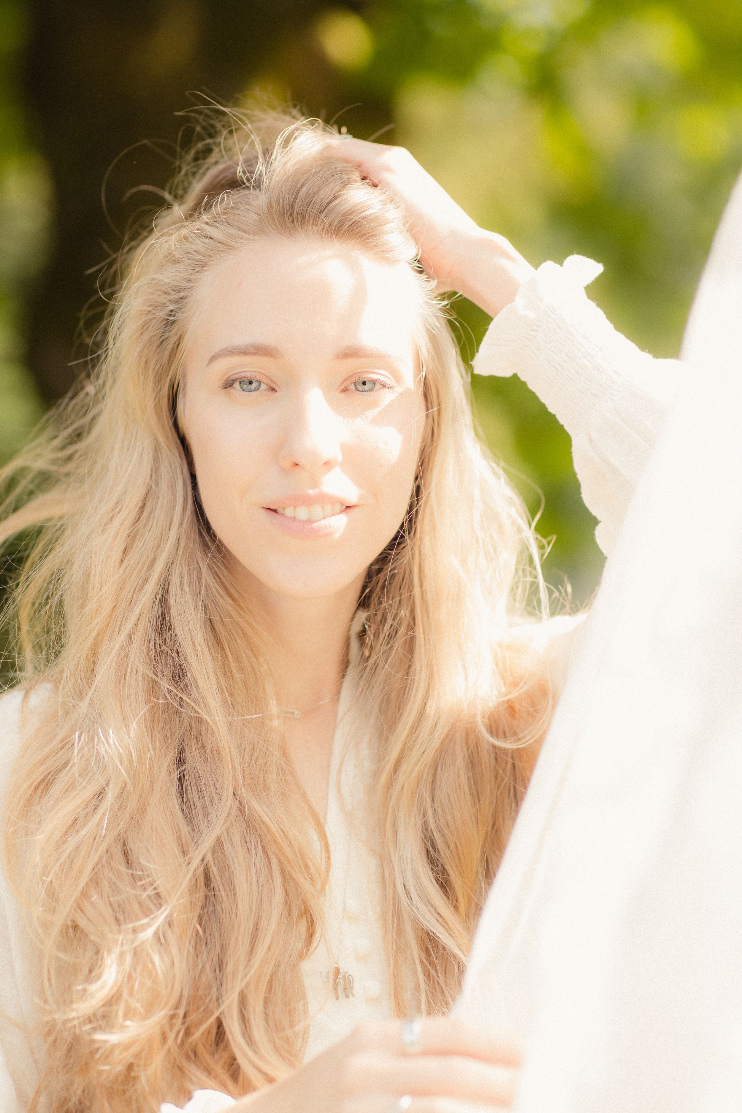Sunlit close-up portrait showing long blonde hair and white sweater against a blurred natural background.