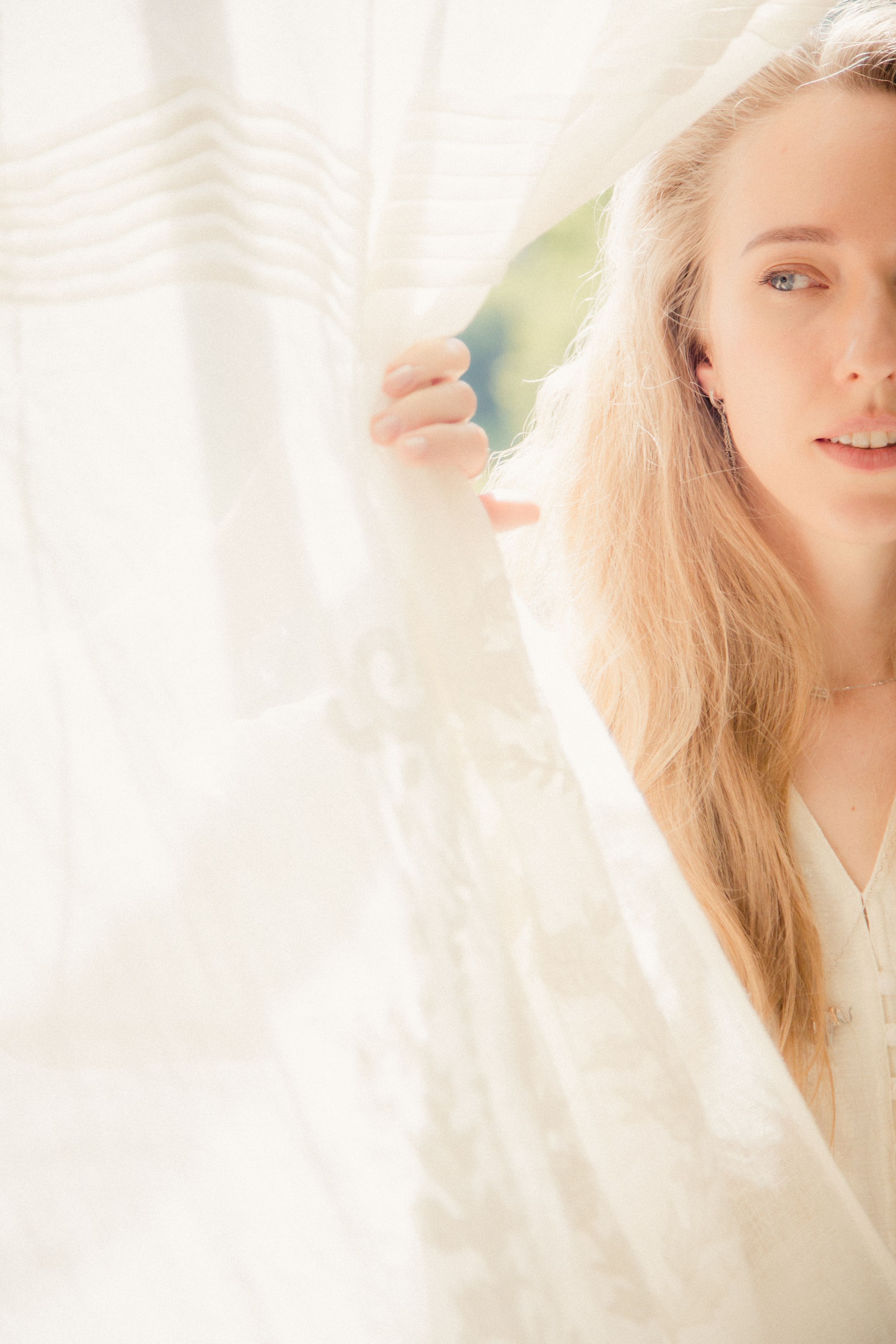 Soft sunlit portrait with sheer white curtains and blonde hair creating an ethereal mood.